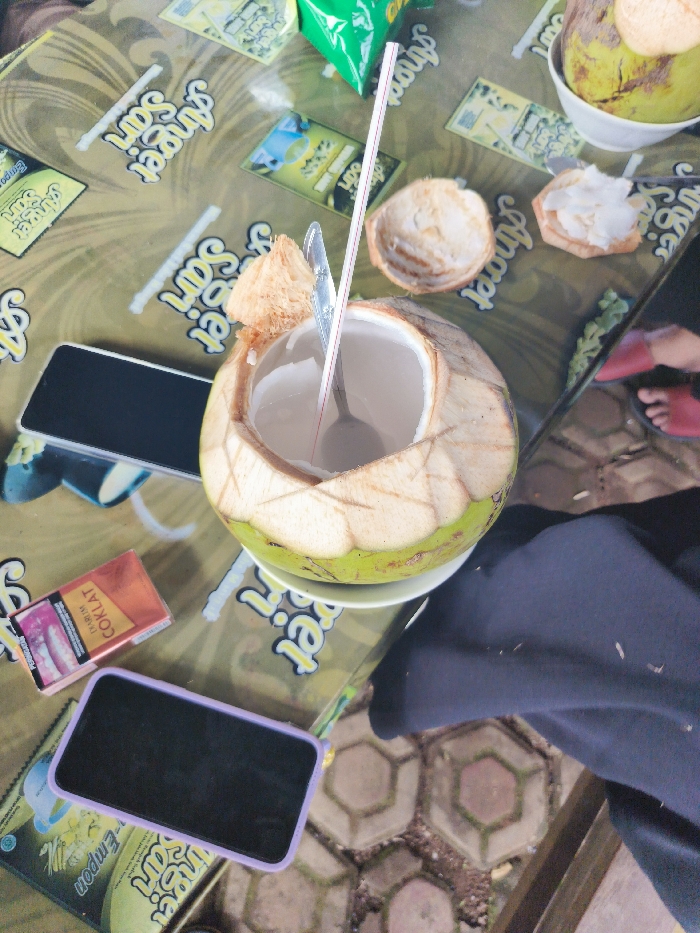 A coconut, cracked open and partially consumed, sits on a table with a drinking straw and spoon inserted. The coconut is surrounded by other coconuts and coconut shells, with a single cigarette pack and two smartphones on the table. The table is covered in a green and yellow tablecloth with the word "Sweet" repeated in multiple locations. The table is situated on a ground covered in hexagonal tiles and a portion of a black garment hangs from the right side of the image. The location is in Majalengka, Indonesia, at coordinates -6.846, 108.204.