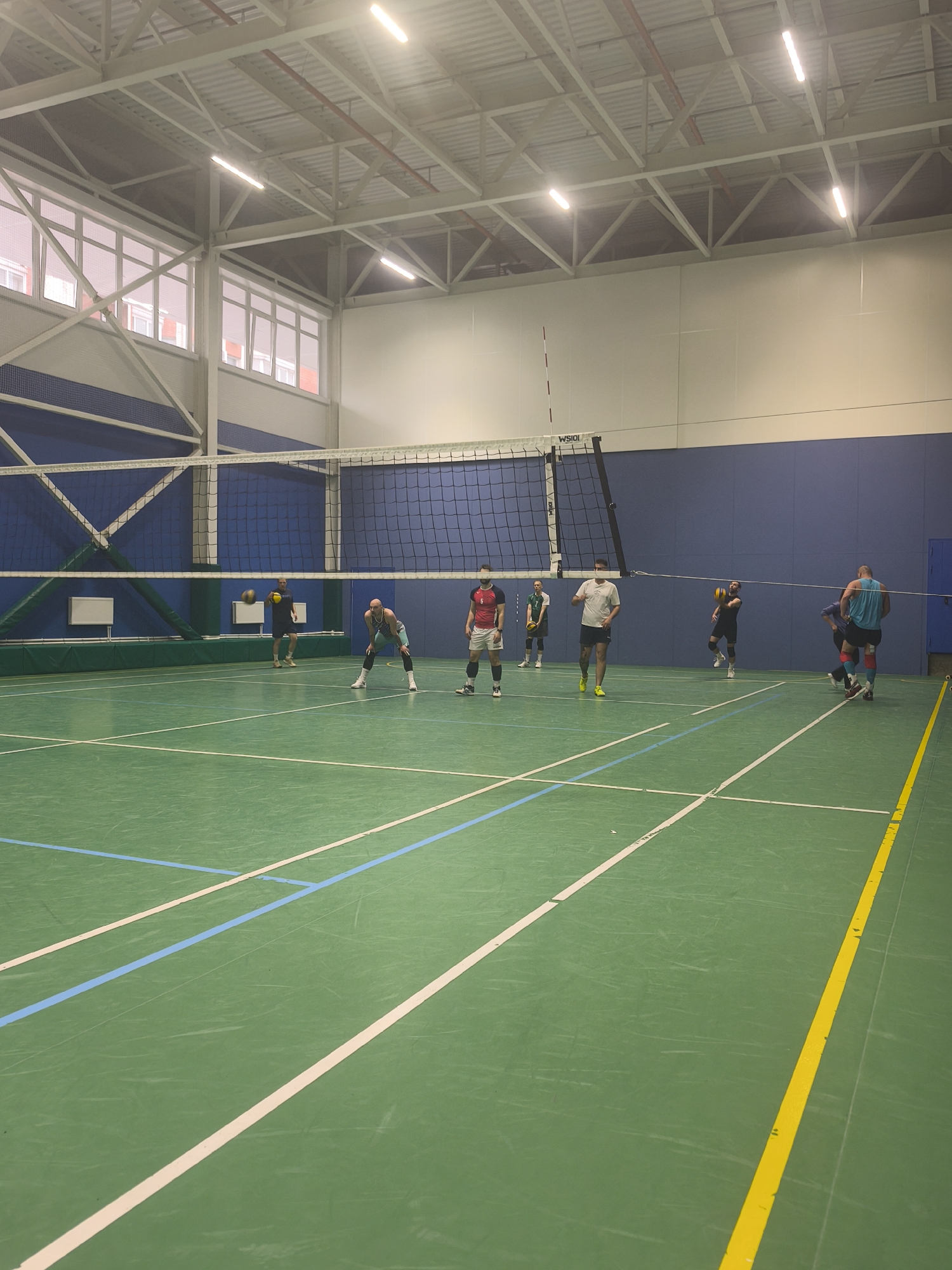 The image shows a group of men playing volleyball indoors, likely in a gymnasium in Saint Petersburg, Russia, given the modern architecture. The court is green with blue and white lines marking the boundaries. A volleyball net stretches across the center of the court. Several men are positioned on both sides of the net, apparently engaged in a volleyball game. Some are wearing athletic attire, including shorts and jerseys. One man is preparing to hit the volleyball, while others are in ready positions. The gymnasium has high ceilings with visible metal support structures. Windows run along one wall, providing natural light. The walls are painted blue and white. The overall environment suggests an active recreational space.