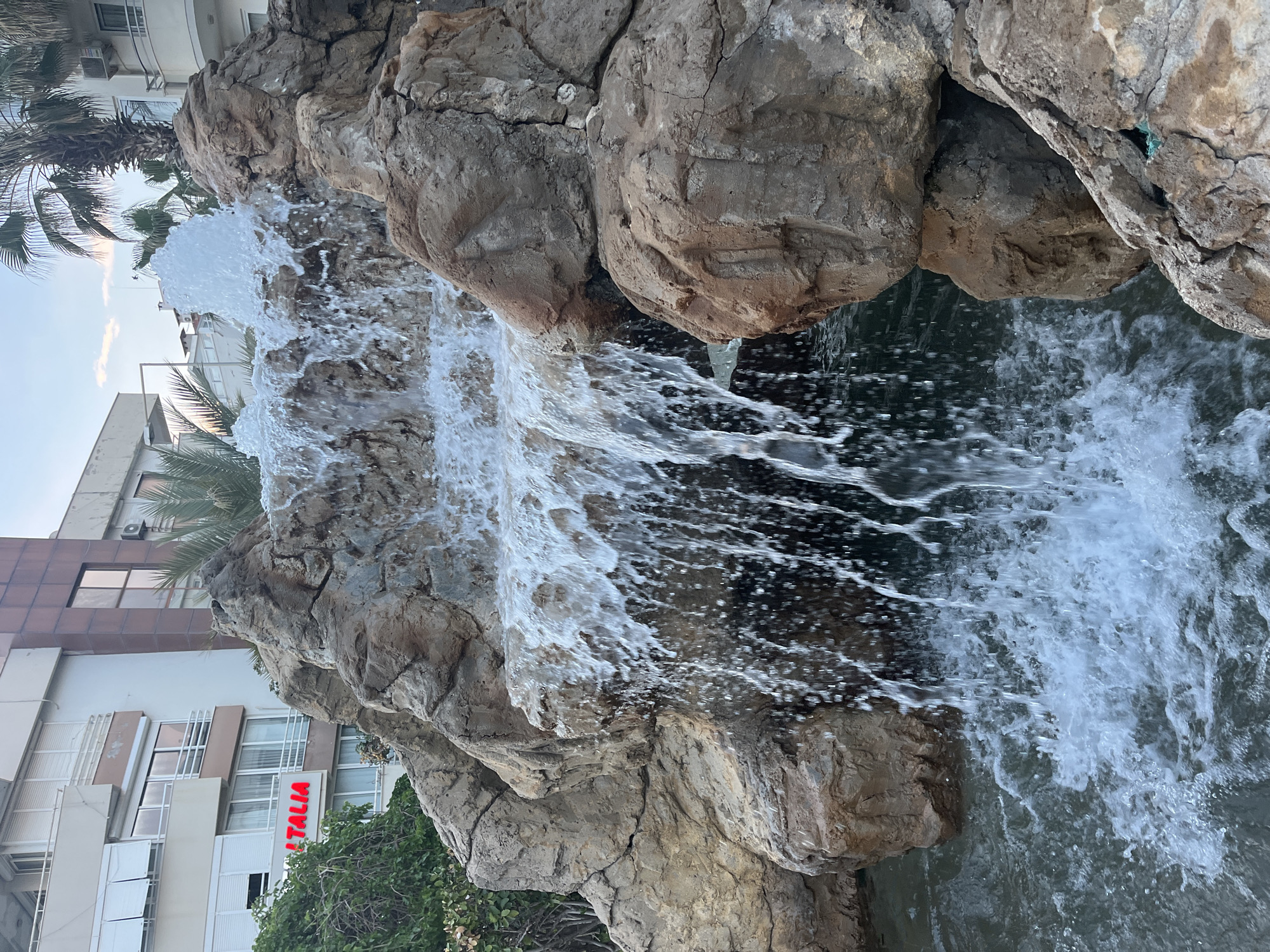 The image captures a vibrant man-made waterfall feature in an urban setting, likely during the daytime. A prominent rock formation, composed of large, textured grey and brown artificial rocks, forms the structure over which water actively cascades and splashes. The water appears white and aerated as it tumbles down, creating dynamic patterns and ripples in a darker pool or basin at the bottom.
To the left and in the upper left background, several palm trees are visible, with their distinctive fronds suggesting a warm climate. Behind the waterfall and foliage, a multi-story building with a modern design is present. It features light-colored walls, numerous windows, and balconies, with some brown and grey architectural details. Air conditioning units are also visible on its exterior.
A significant detail on the building is a red vertical sign displaying the word "ITALIA." Green bushes and other foliage are visible at the base of the rock formation and in front of parts of the building. The sky, partially visible above the building and through the palm trees, is a clear light blue, indicating fair weather. The scene depicts an attractive water feature integrated into an urban environment, consistent with a location like Alanya, Turkey.
