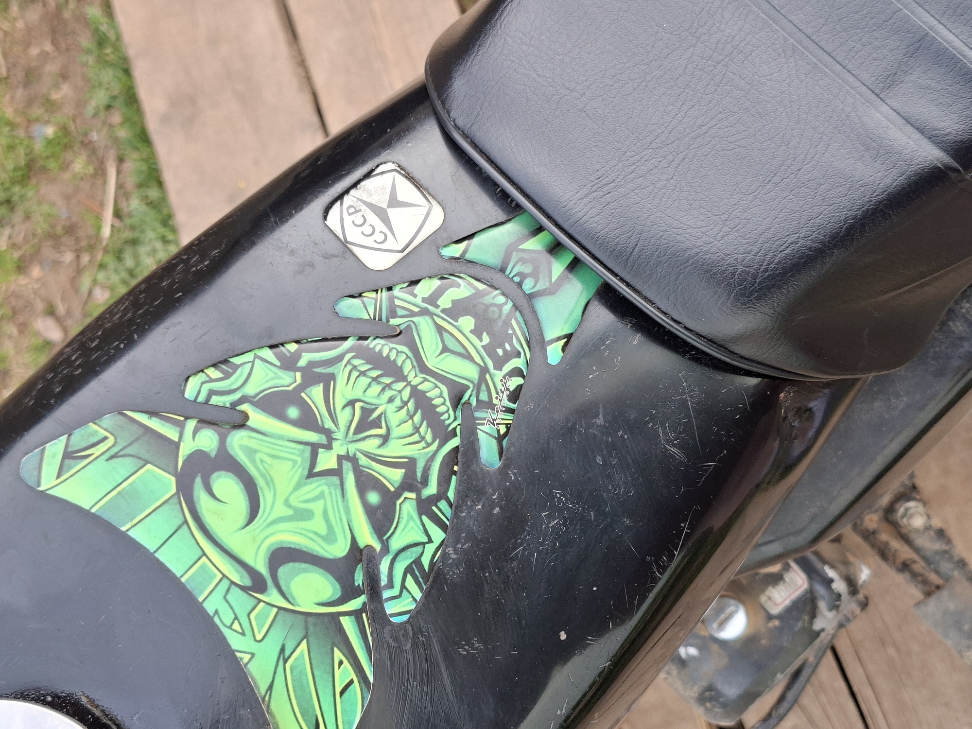 A close-up photograph depicts a section of a black motorcycle, likely outdoors, with a focus on the rear portion near the seat. The motorcycle features a green and black patterned decal on the body, partially obscured by the black leather seat. A square sticker with "CCCP" written in Cyrillic alphabet is affixed near the decal. The surface of the motorcycle shows signs of wear, with scratches visible on the black paint. The background includes a wooden surface, possibly a platform or ground cover, suggesting an informal outdoor setting.