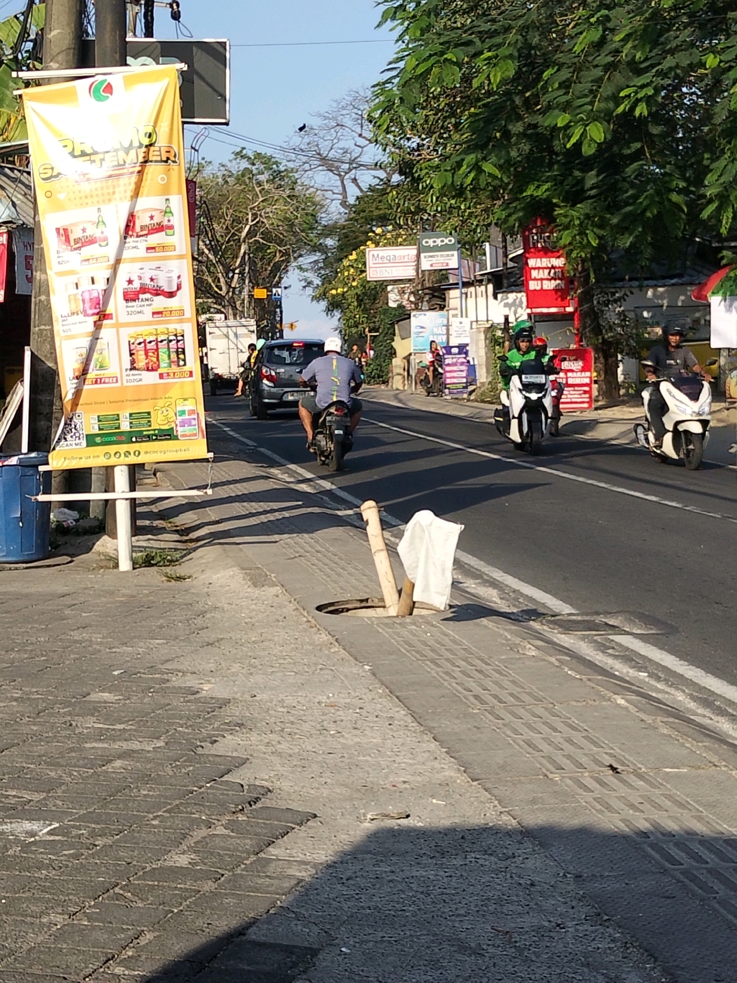 The image depicts a street scene in Kuta Selatan, Indonesia, characterized by a sunny day and bright lighting. The foreground shows a cracked and uneven sidewalk with paving stones. A bamboo pole with a white cloth tied to it stands near a manhole cover. In the middle ground, a paved road is visible with white lane markings. Several people are on motorcycles; one man in a gray shirt and helmet is riding a motorcycle and appears to be looking towards the right. Another motorcycle rider in a green jacket and helmet is also on the road. A silver car is seen in the background, traveling away from the viewer. A large yellow banner advertising "PROMO SEPTEMBER" is prominently displayed on the left side of the image, featuring images of beverages and snacks, along with prices. Various signs for businesses like "Megaponto" and "oppo" are also visible. The environment includes trees with green foliage and a bright blue sky. The overall impression is a busy urban street scene in a tropical climate.