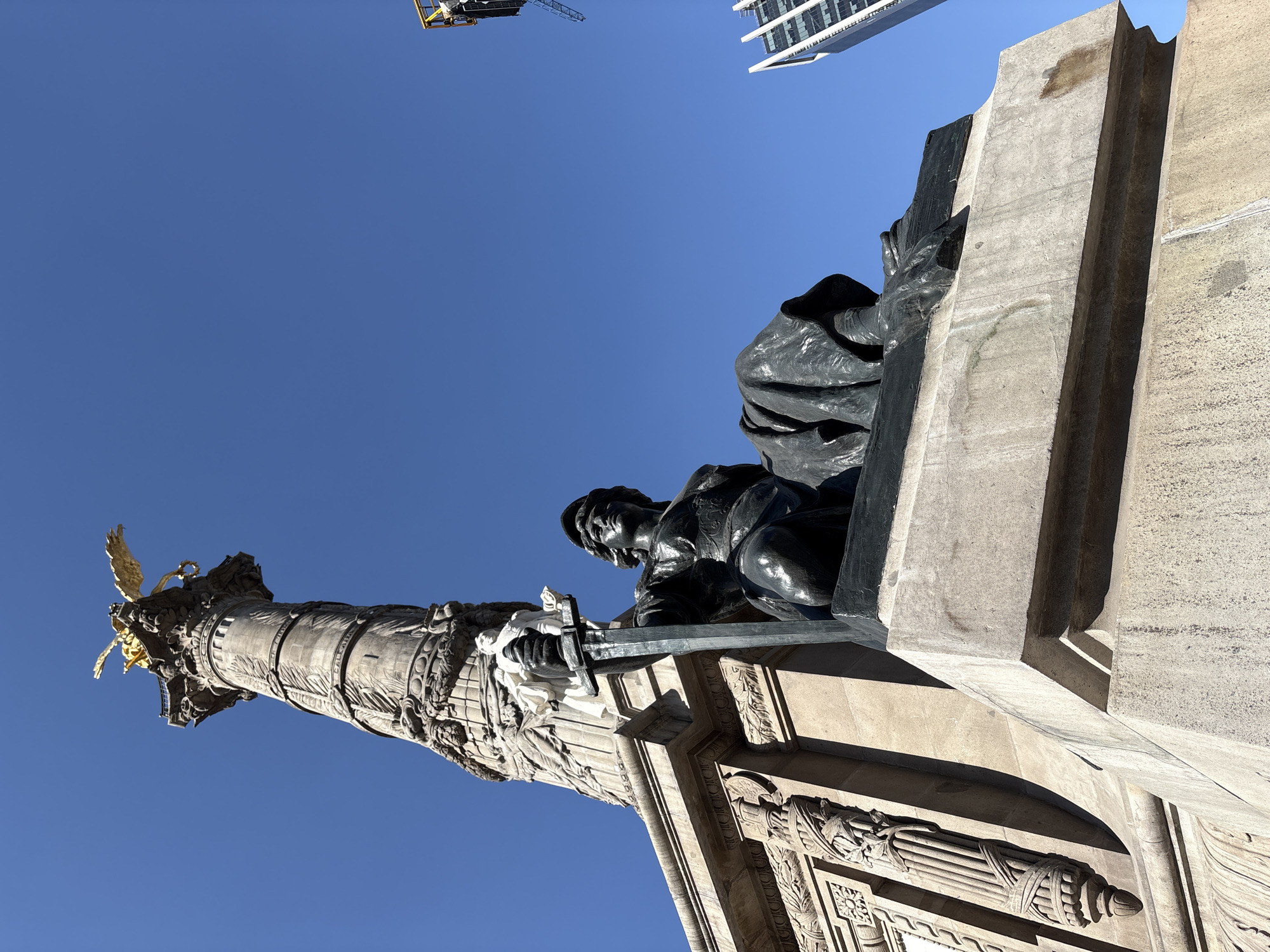 An upward-angled outdoor photograph captures a portion of the Monumento a la Independencia, also known as El Ángel de la Independencia, located on Paseo de la Reforma in Mexico City, Mexico. The central subject is a tall, ornate stone column surmounted by a golden winged figure.
In the foreground, a dark bronze allegorical statue sits upon a light-colored stone plinth. This figure, depicted with a helmet and draped garment, holds a large sword vertically by its hilt with both hands, the blade pointing upwards. The stone plinth exhibits minor discoloration and weathering.
The main column of the monument is constructed from light-colored stone and features extensive carved reliefs, including floral garlands and bundled motifs resembling fasces. Further up the column, lighter-colored statues are recessed into the structure. The column is crowned by a golden, winged victory figure.
The sky is uniformly clear and bright blue, indicative of daylight. In the upper-right background, a modern high-rise building with a glass and white panel facade is partially visible, along with the yellow boom and dark structural components of a construction crane. The scene is brightly illuminated by direct sunlight, creating distinct shadows and highlighting textural details.