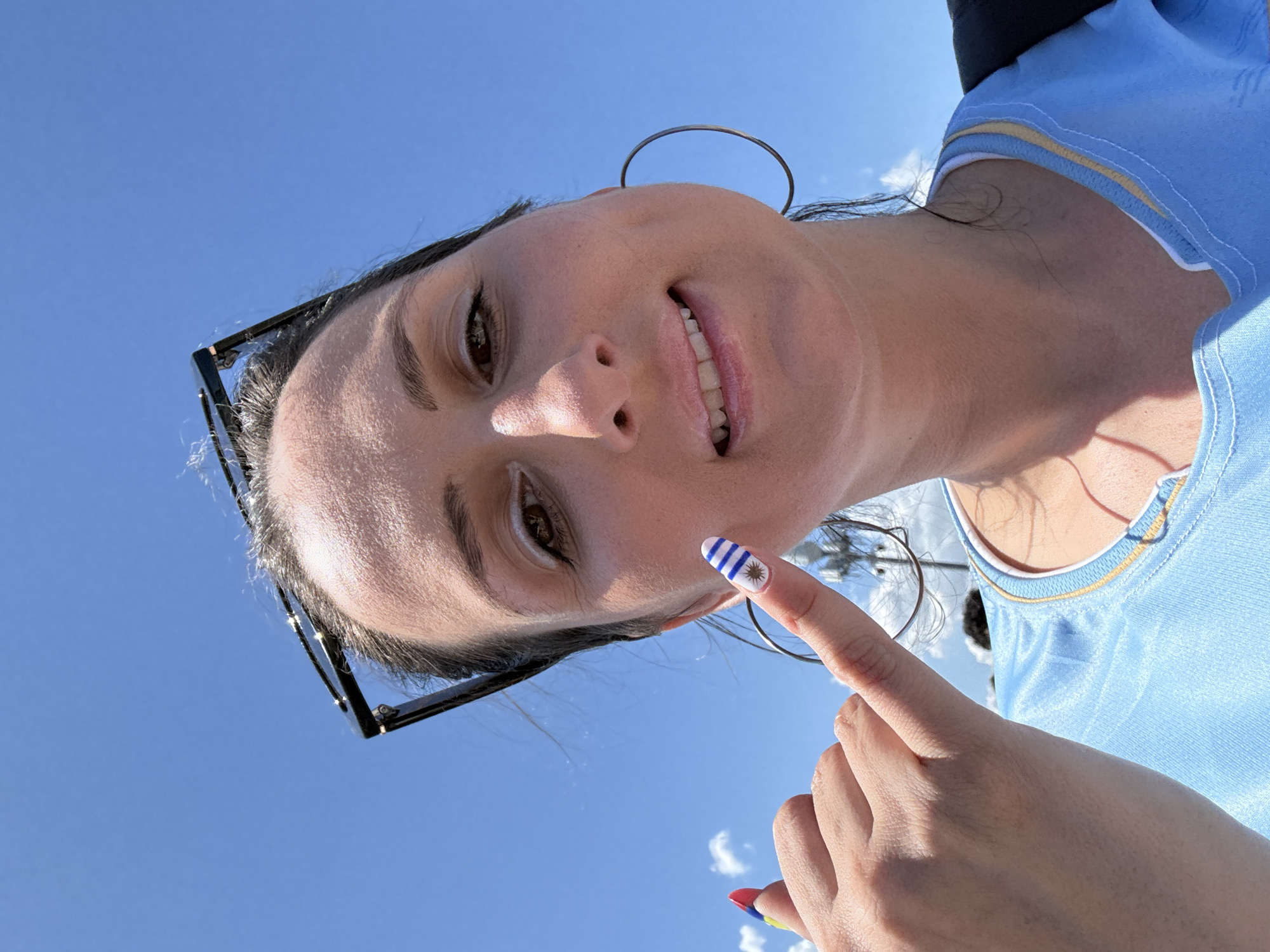 A woman in Miami Gardens, Florida, smiles at the camera while holding up her right index finger, which is painted with the flag of Uruguay. The flag design features white and blue stripes with a golden sun in the canton. The woman wears a light blue t-shirt with yellow and white trim. She has on large hoop earrings and sunglasses perched on her head. The background is a clear blue sky.