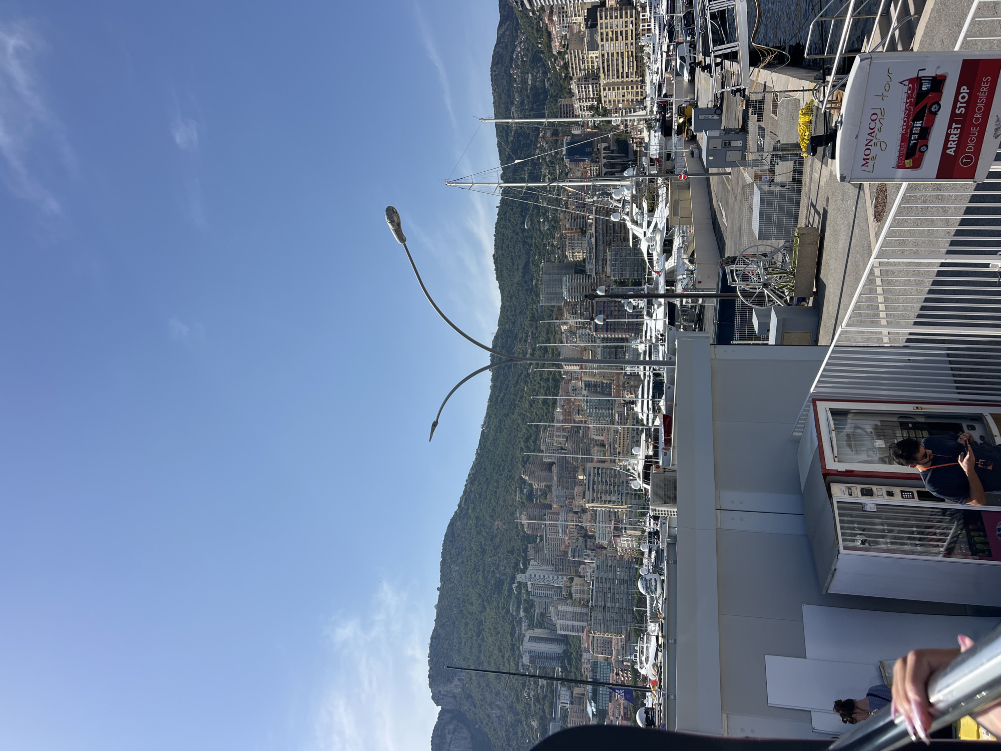 This is a wide-angle, eye-level shot of a harbor in Monaco, with a cityscape rising behind it. The sky is clear and blue with a few wispy clouds. In the foreground, on the left, a person's hand with long, manicured nails is visible, gripping a railing. To the right, a vending machine stands next to a man wearing a dark t-shirt and sunglasses, looking at his phone. Behind them, a white railing separates the viewer from a road and a harbor filled with numerous yachts of varying sizes. The city of Monaco is built on the slopes of a verdant hill that dominates the background. The architecture is dense, with many tall buildings clustered together. The image captures a sunny day in a bustling Mediterranean port.