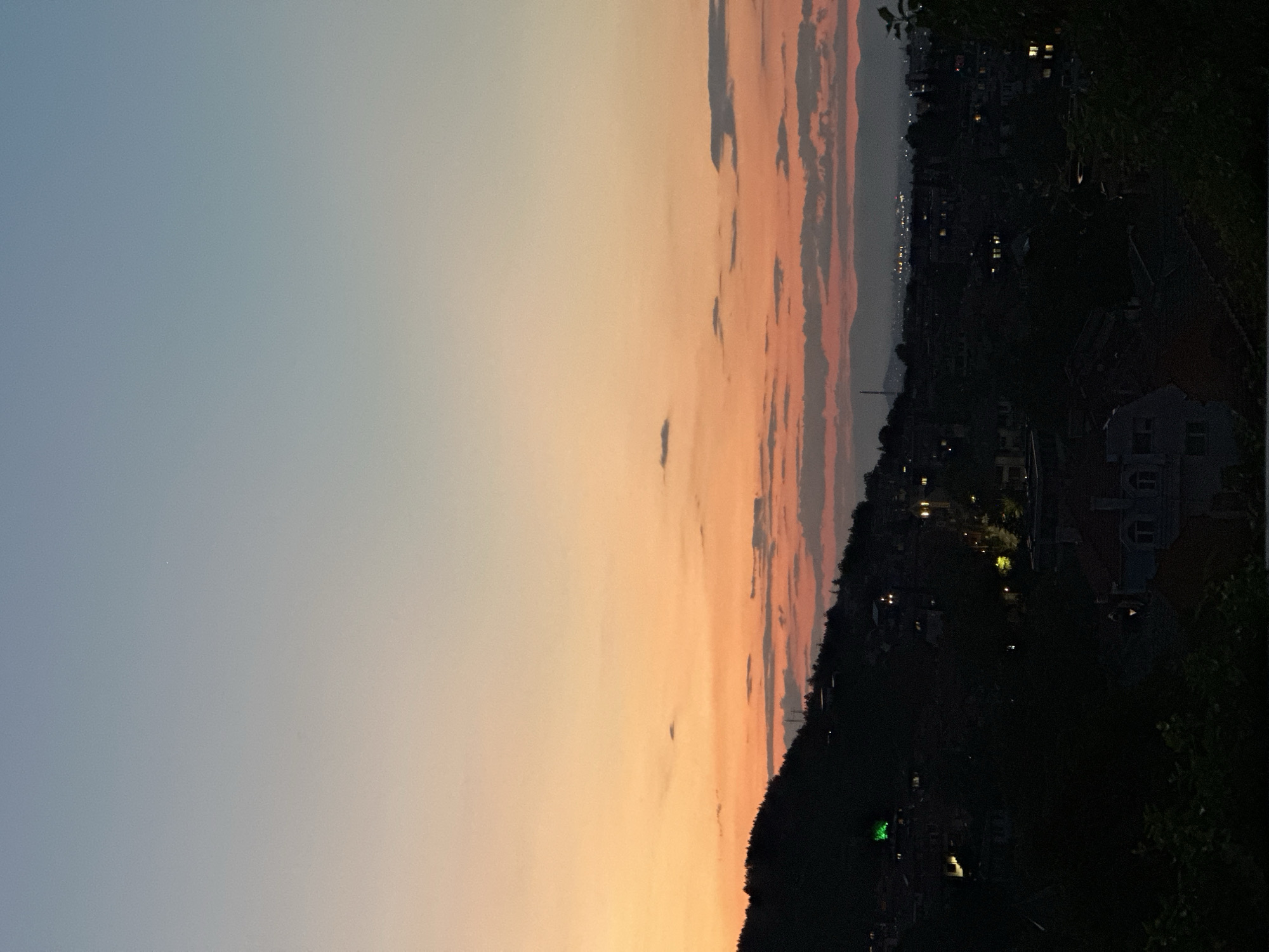 The image displays a panoramic view of Asenovgrad, Bulgaria, at dusk from an elevated perspective. The sky exhibits a gradient, transitioning from a light blue at the top to a vibrant peach and orange band along the horizon, with several horizontally elongated dark grey clouds. A singular bright white point of light, likely a star or planet, is visible in the upper right quadrant of the sky.
A dark, heavily treed hillside dominates the left midground, sloping downward towards the right. A distinct, bright green light source is visible within the foliage on this hillside, and a slender, dark mast-like structure is silhouetted against the sky at its peak. Below the hillside and extending into the midground and foreground, the urban landscape of Asenovgrad is visible. Buildings are largely obscured by encroaching darkness and shadows from trees and topography. Numerous artificial lights from residences and streetlamps illuminate portions of the city, appearing as warm yellow and orange points of light.
In the far background, a hazy, dark mountain range is silhouetted against the horizon, with a more densely lit urban area extending beneath it, indicating a broader expanse of the city or a distant settlement. No individuals or specific actions are discernible within the frame.