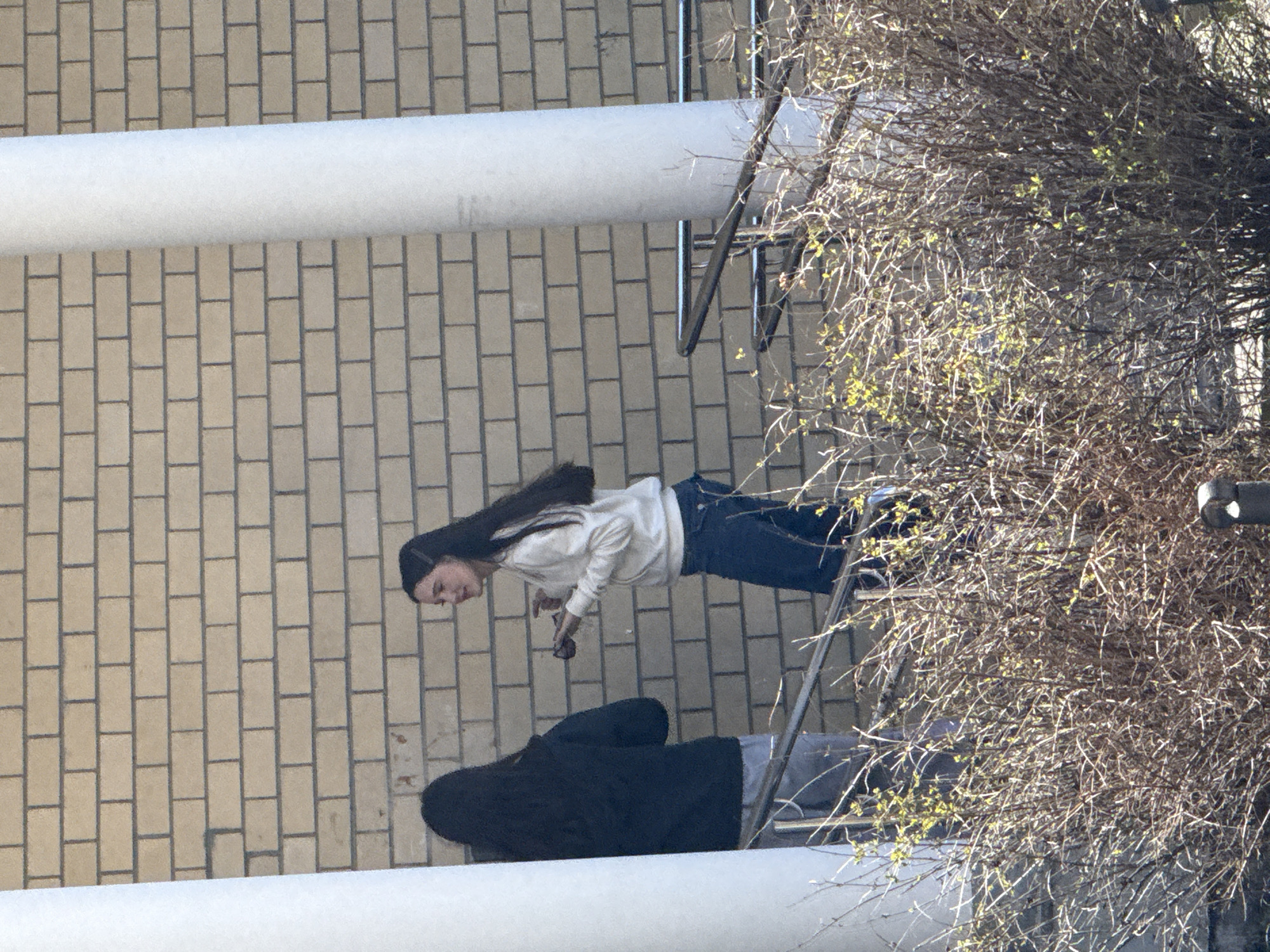 A high-angle outdoor shot depicts a woman with long, dark hair and sunglasses, wearing a white sweatshirt and blue jeans, standing on a balcony. To her left, another individual in a dark hoodie and gray pants reclines on the balcony. A brick wall is visible in the background, along with metal railings and a bare tree branch. The location is potentially Yekaterinburg, Russia.