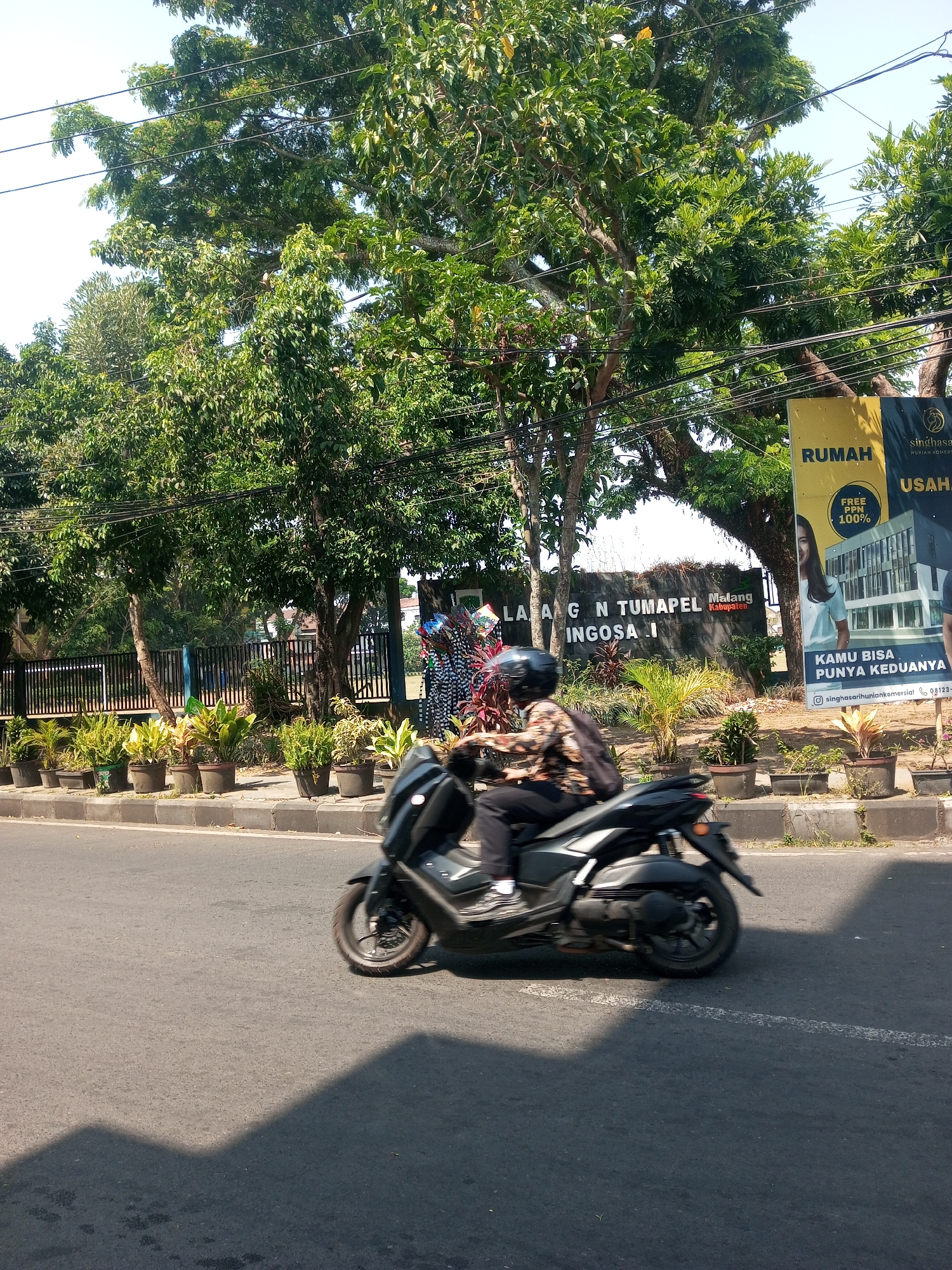 A person wearing a helmet rides a black motorcycle on a paved road. The rider is wearing dark pants and a patterned shirt. In the background, there are large trees, a black fence, and a sign that reads "LABANG N TUMAPEL Malang Kabupaten". There are also potted plants along the side of the road and a yellow banner with text in Indonesian. The sky is clear and the scene appears to be in Singosari, Indonesia, as indicated by the sign.