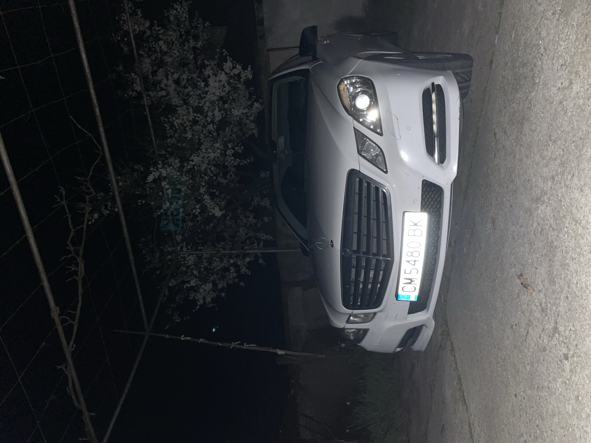 The image shows a white Mercedes-Benz car parked on an asphalt surface, potentially a road or driveway. The license plate number is CM 5480 BK. The environment appears to be a residential area or edge of a town, suggested by a blooming tree and a wire fence in the background. It is nighttime, with limited artificial light sources illuminating the car. The location is reported to be Zlatograd, Bulgaria. There are no people visible in the image.