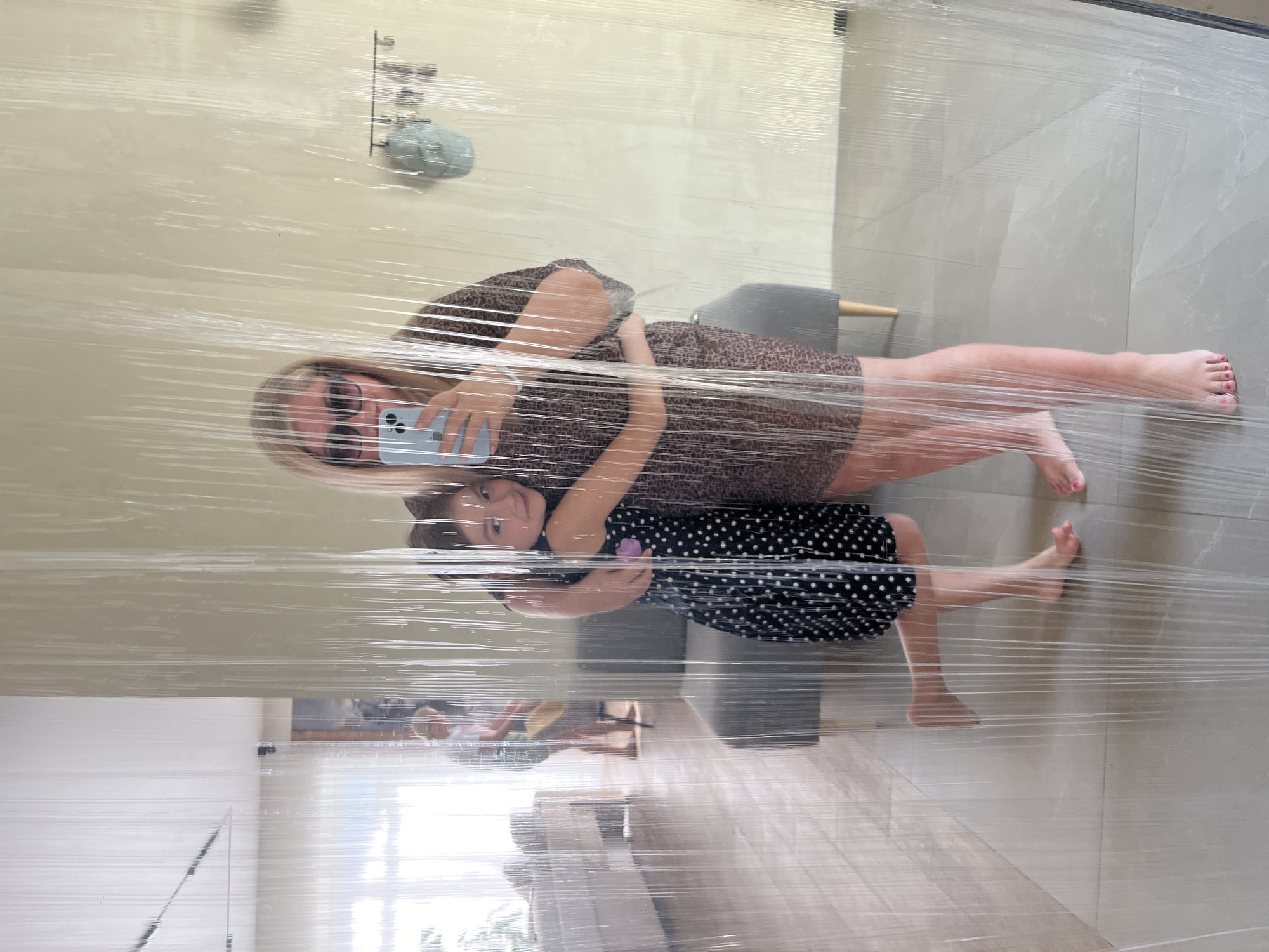 A woman and a child are pressed against a glass surface, likely a window or mirror, wrapped with clear plastic film. The woman is wearing a brown patterned dress and sunglasses, holding a blue smartphone, presumably taking a photo. The child is wearing a black dress with white polka dots. The background includes a tiled wall, a chair, and a partially visible interior of a room with furniture and a window. The location is identified as Новая Чернушка, Russia.