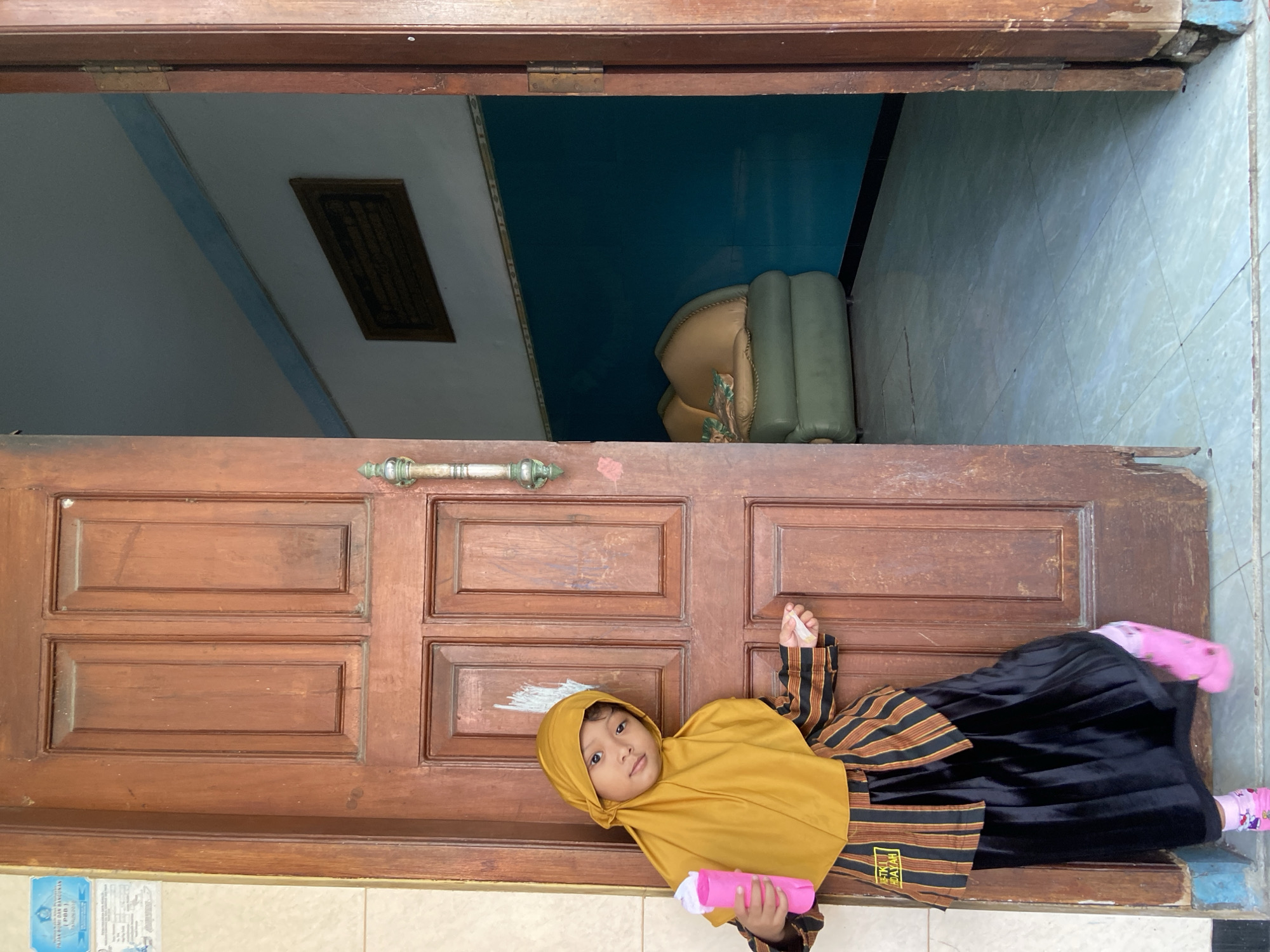 A child is lying across an open wooden doorway. The child wears a mustard-colored hijab, a black and brown striped dress, and pink socks. She holds a pink object wrapped in white cloth. Inside the doorway, a room is visible with blue-painted walls and tiled floors. A tan and green chair is visible within the room. The location is Gelam, Indonesia.