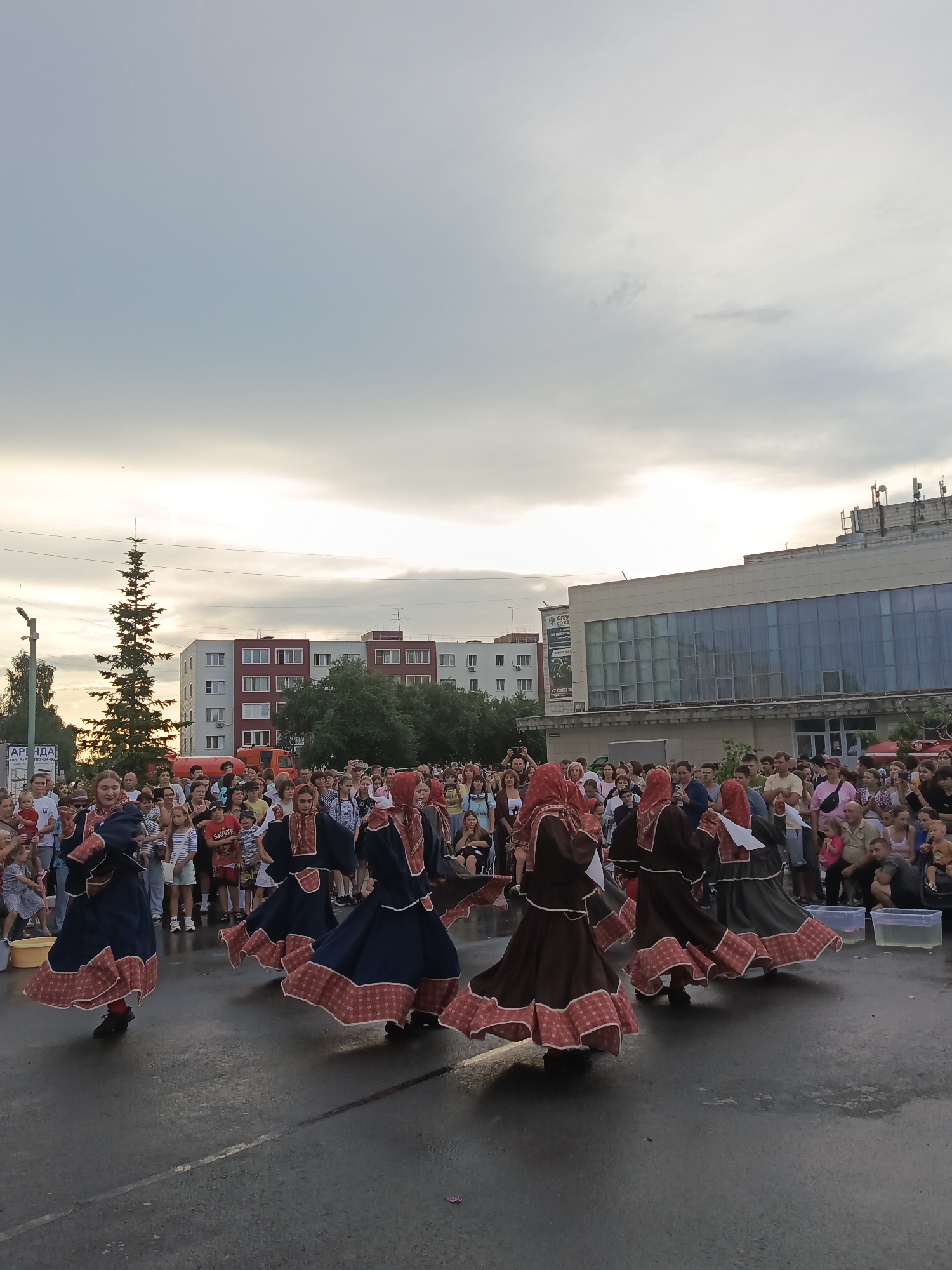 In this wide shot, a group of people are gathered outdoors, possibly for a festival or cultural event. The main subjects are several young women in traditional folk costumes, characterized by long, flowing dresses with wide skirts and decorative trim. They are dancing in a circular formation on a paved surface that appears slightly wet. The dancers are adorned in dresses of blue and brown, with red and white patterned borders on their hems and sleeves, and matching headscarves. They are facing away from the camera, their movements creating a sense of motion.
Behind the dancers, a diverse crowd of spectators is visible, many standing and observing the performance. The spectators are dressed in casual, modern clothing. In the background, there are multi-story buildings, one with a modern facade featuring large, reflective windows, and another with a more traditional brick design. A tall, slender evergreen tree stands to the left, adding to the outdoor setting. A fire truck is also partially visible among the buildings.
The time of day appears to be late afternoon or early evening, as indicated by the soft, diffused light and the overcast sky, with hints of sunlight breaking through the clouds. The atmosphere seems festive and engaging, with the audience attentively watching the performance. No specific emotions are overtly displayed by the crowd, but the scene suggests a lively community gathering.
Visible text includes a sign on a pole in the foreground on the left, partially obscured, but the letters "АРЕНДА" (ARENDA, meaning "rent" or "lease" in Russian) are discernible. A sign on the building in the background also displays some text and graphics, but it is too small to be clearly read. The overall setting suggests a public square or plaza in a town or city in Russia.