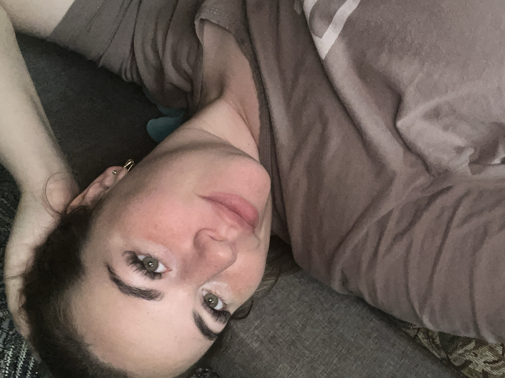 A woman is lying down on a couch, with her head propped up by her hand. She is wearing a brown t-shirt. She has long eyelashes, and her hair is dark brown. The background is a couch with a patterned blanket. The image appears to be taken indoors. There is no information that would suggest this image is located in Chaikovsky, Russia.