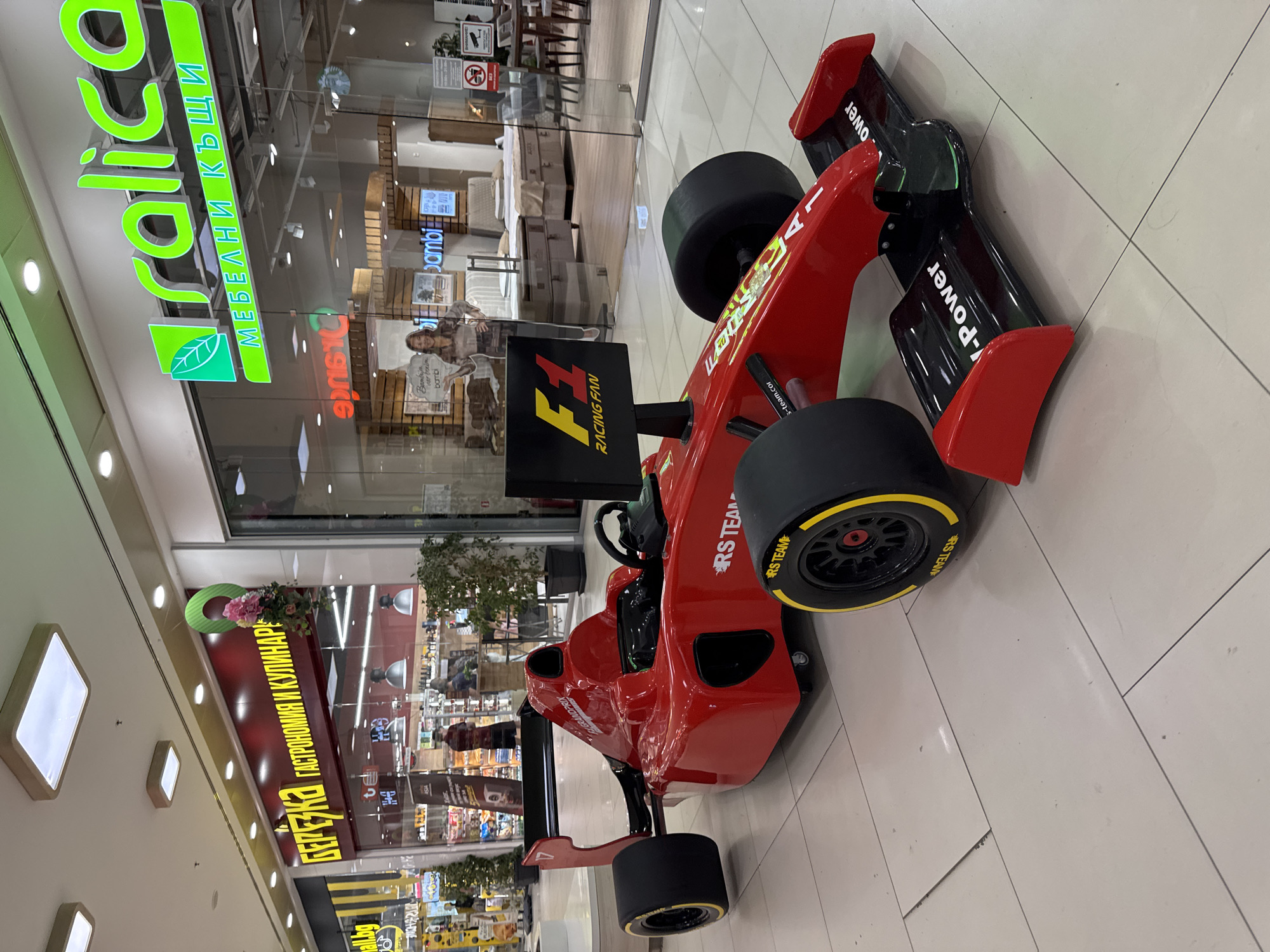 This image depicts an indoor scene within a shopping mall in Plovdiv, Bulgaria, specifically identifiable as likely *Mall Plovdiv* due to the presence of the "БЕРЁЗКА" (Berezka Gastronomy and Culinary) and "Sralica" furniture stores.
In the central foreground, a large, red, two-seater display model of a Formula 1 racing car is positioned on a light grey tiled floor. The car features black tires with yellow "RS TEAM" branding, "F1 GRAND PRIX" and "RS TEAM" logos on its red bodywork, and "V-Power" inscribed in white on its black front wing. A black sign attached to the front of the vehicle displays "F1 RACING FAN" in yellow and red text.
To the left, the storefront of "БЕРЁЗКА ГАСТРОНОМИЯ И КУЛИНАР" (Berezka Gastronomy and Culinary) is visible, characterized by yellow Cyrillic lettering on a red background. A green, circular decorative element adorned with pink artificial flowers hangs near the store sign. Through the glass, shelves displaying various products are visible, along with two individuals inside; one male figure in a reddish top is partially obscured.
To the right, the storefront for "Sralica МЕБЕЛНИ КЪЩИ" (Sralica Furniture Houses) is present, featuring bright green lettering and a leaf logo. Interior displays visible through the glass include multiple beds with various bed frames and mattresses, and a dining set. A life-sized cardboard cutout of a woman stands next to a "bambi" branded display. A red "NO SMOKING" pictogram and a "ВИДЕОНАБЛЮДЕНИЕ" (CCTV) warning sign are affixed to the right-hand glass door. A faint reflection of a Starbucks logo is visible on the glass of the Sralica store.
The overall environment features a polished, light-colored tiled floor with visible grout lines and reflections. The white ceiling is equipped with a variety of flush-mounted rectangular and circular lighting fixtures.