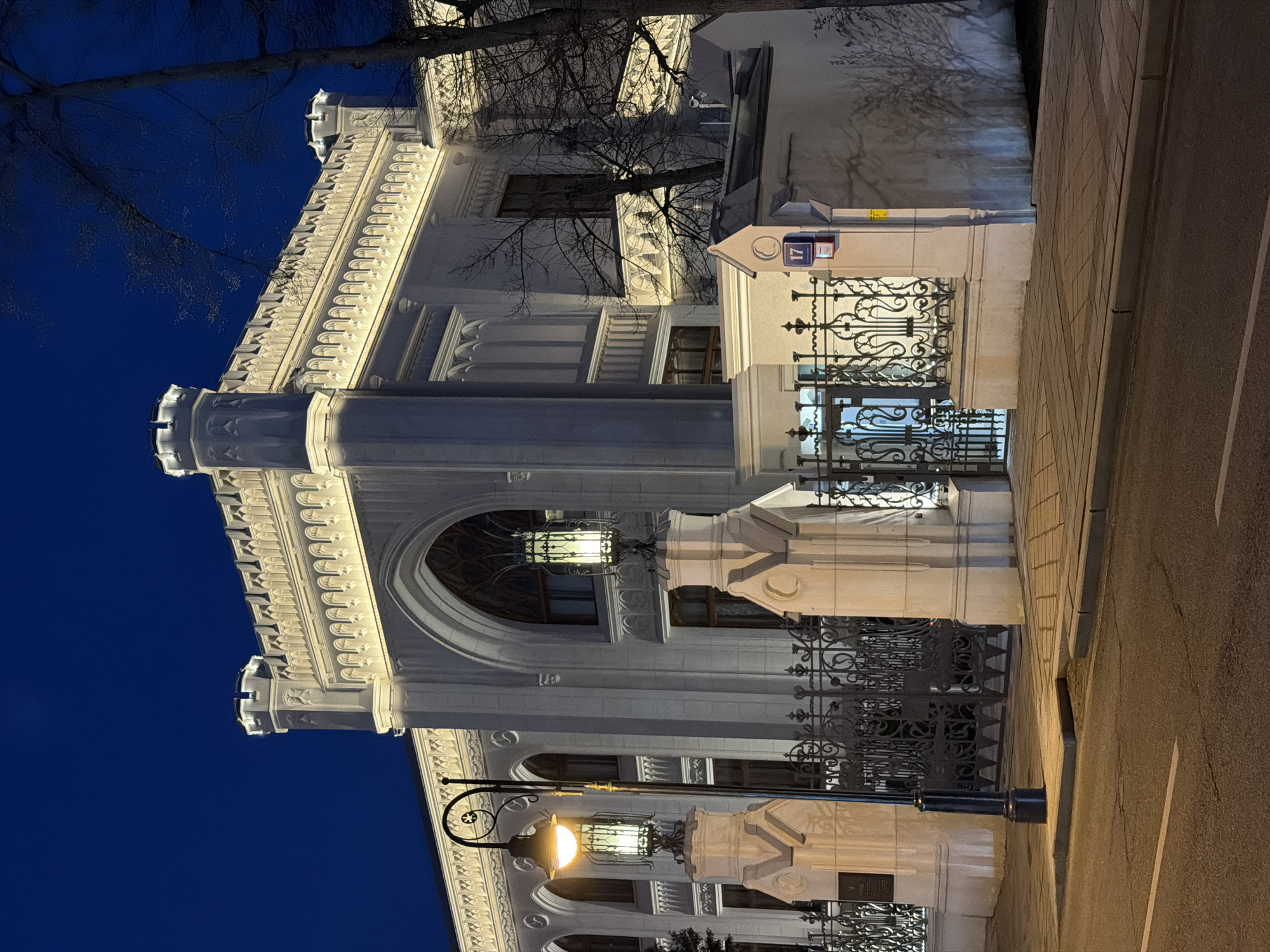 The image shows a section of the Perl House at night, located at Chistoprudny Boulevard, 17 in Moscow, Russia. The building features elaborate architectural details including arched windows, decorative moldings, columns, and ornate wrought iron gates. A street lamp illuminates a section of the building's facade. A tree with bare branches is visible near the top of the building. The sky is dark blue. The street is visible on the right side of the image.