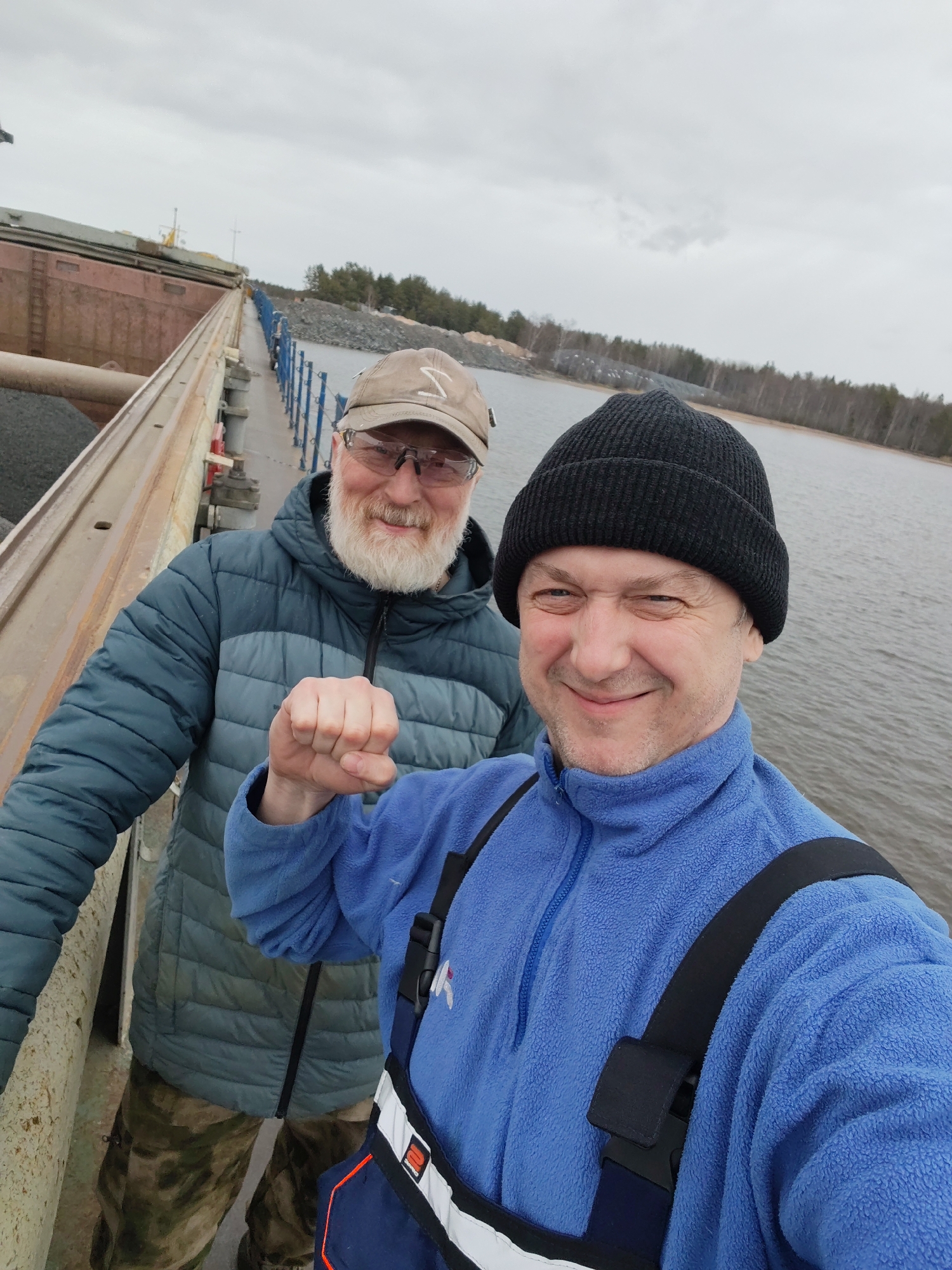 A selfie captures two smiling men standing on a pier or walkway beside a large vessel under an overcast sky, likely in Shcheleiki, Russia.
The man on the left, appearing older, has a full white beard, wears glasses, a light brown baseball cap with an indistinct emblem, a blue and grey puffer jacket, and camouflage patterned trousers. He is smiling warmly.
The man on the right, who is taking the selfie, wears a black knitted beanie, a vibrant blue fleece zip-up jacket, and dark blue work overalls with black shoulder straps and white and orange accents. He smiles broadly and holds up a clenched fist in a friendly or enthusiastic gesture.
Behind them, a large, weathered, reddish-brown barge or ship is moored, with a portion of its cargo, resembling gravel or crushed stone, visible. A narrow pier or walkway with blue railings extends into the background, separating the vessel from the open water. The distant shore features a dense line of trees, typical of a northern forest, and industrial piles of material, suggesting a working port or quarry area. The sky is completely clouded, indicating cool weather, which is consistent with the men's heavy clothing. Both individuals convey a cheerful and positive mood. No discernible text or specific time of day can be clearly identified.