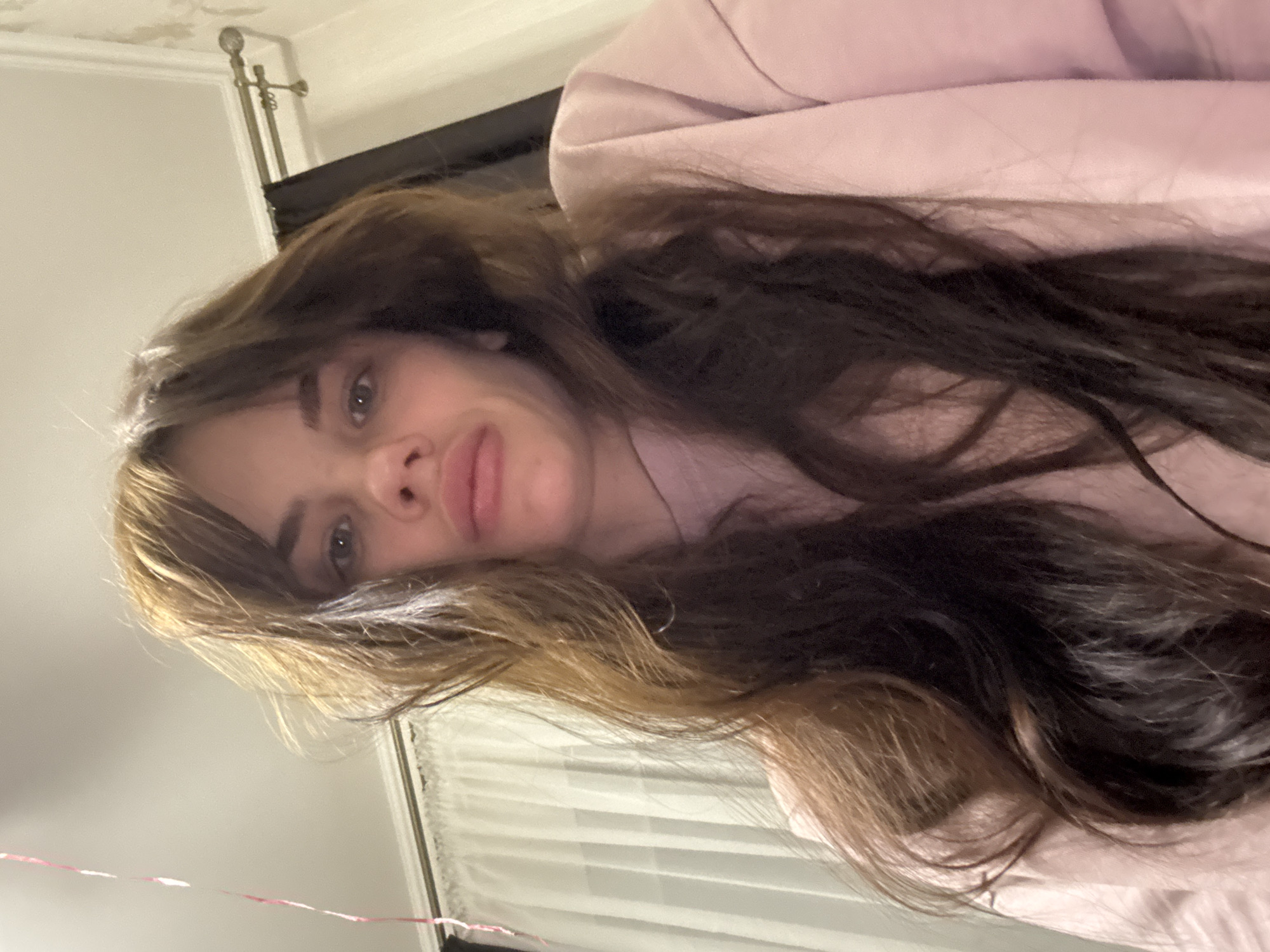 A selfie photo depicts a woman indoors. Her hair is long, a mix of dark brown and lighter brown/blonde tones, framing her face. She is wearing a light pink top. Behind her are window blinds and a headboard. A wall and molding are partially visible in the background. There is no information to indicate that the location is Rostov-on-Don, Russia.