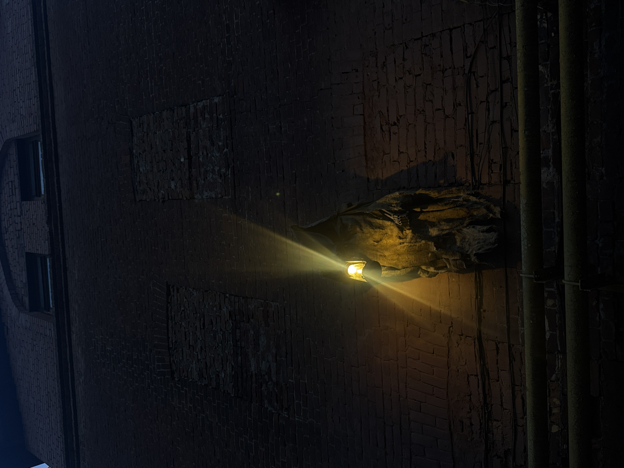 An outdoor low-angle shot shows the side of a brick building at night. The building is dark red and has filled-in windows and metal pipes running vertically on the right side. On the center right, a light fixture is attached to the wall, providing illumination. The light shines brightly, creating a dramatic effect. The area around the light fixture appears weathered and textured. The overall scene conveys an urban, possibly industrial, environment. There is no indication of people in the image.
