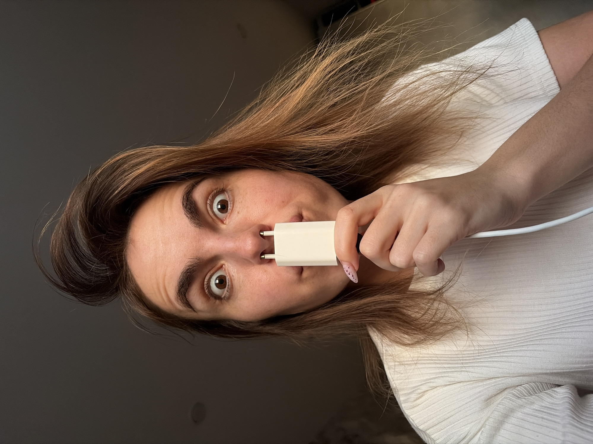 A woman with brown hair is holding a white European-style electrical plug to her face, inserting the prongs into her nostrils. She is wearing a white ribbed shirt. The background is a gray wall. The image is rotated counter-clockwise. There is no indication of the location being Izhevsk, Russia.