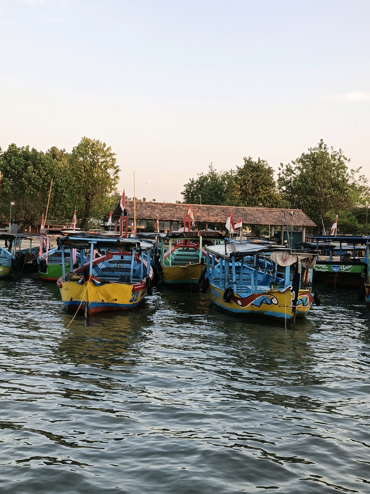 A group of colorful fishing boats are docked on the water in the city of Cikal, Indonesia. The boats are painted in vibrant colors such as yellow, blue, and green, with some featuring decorative artwork. Several boats have red and white Indonesian flags attached to their poles. In the background, there is a building with a tiled roof, surrounded by trees. The water is slightly rippled, reflecting the colors of the boats and the sky. The scene appears to be captured during a calm day, possibly in the late afternoon or early evening, given the soft lighting.