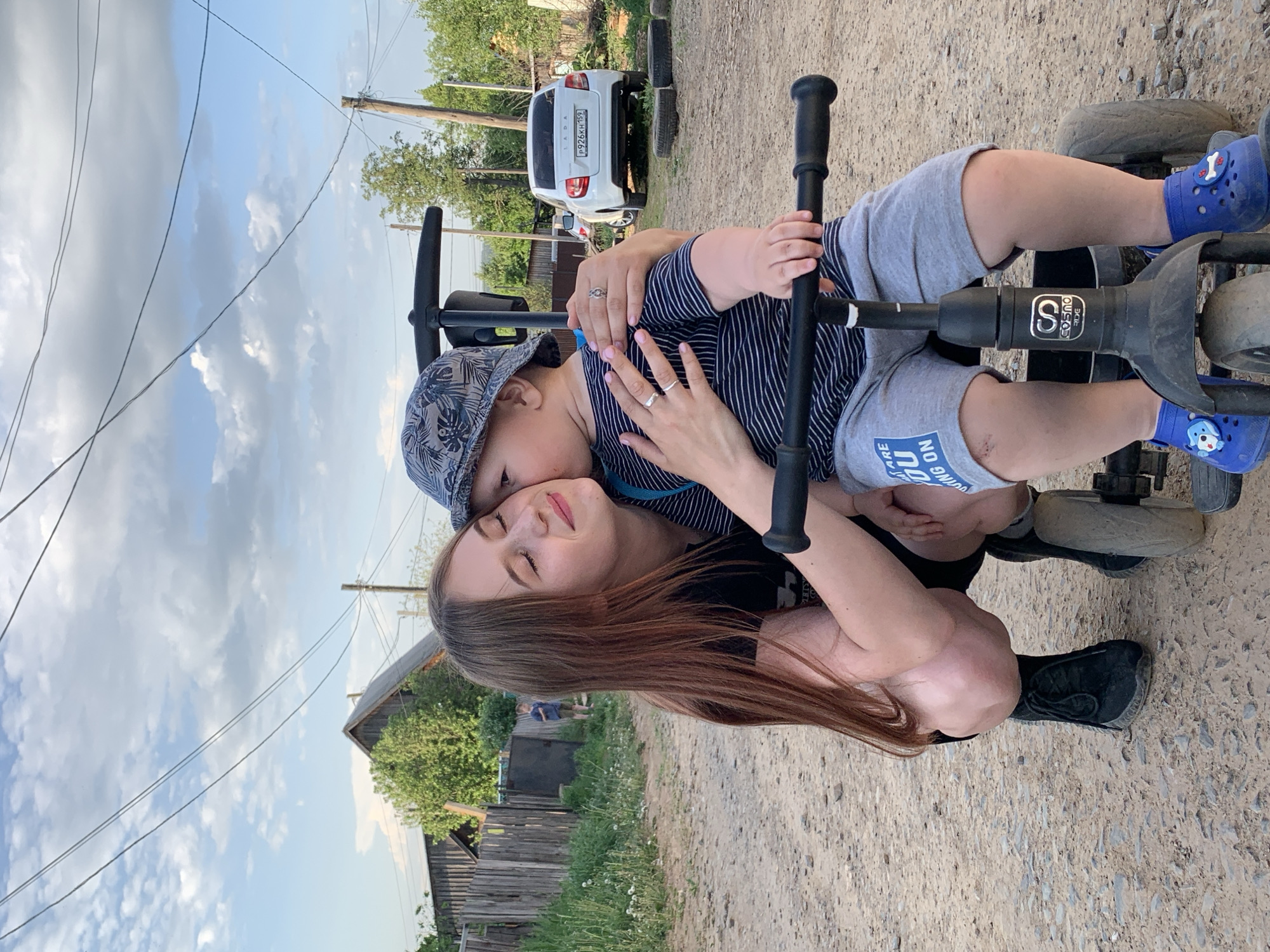 The image shows a woman and a child on a tricycle in a rural or semi-rural environment. The woman is kneeling and hugging the child, who is seated on a black tricycle. The child wears a hat, a striped shirt, gray shorts, and blue shoes with cartoon characters. The woman has long brown hair and wears a black top. Her eyes are closed and she appears to be at peace. Her ring on her finger indicates that she is married. In the background, there is a white car, wooden fences, trees, power lines, and a cloudy sky. The setting appears to be a dirt or gravel road. According to the license plate on the car, the picture was taken in Фоки, Russia.