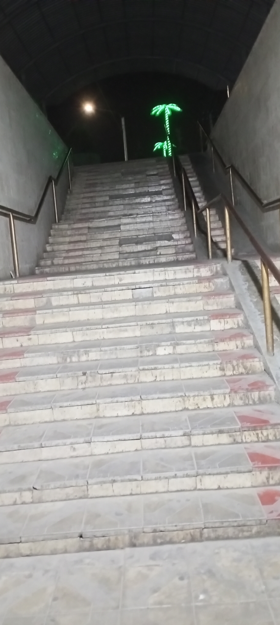 This is a low-angle, full-shot photograph of a wide, outdoor staircase in Sokuluk, Kyrgyzstan, at night. The staircase is constructed from light-colored stone or concrete, with some sections showing signs of wear and tear, including red tiling on some risers. Metal handrails run along both sides of the stairs. The staircase leads upwards into a dimly lit tunnel or covered walkway. Above the top of the stairs, two illuminated green neon palm tree sculptures are visible against a dark background. A single, bright street light is also present to the left of the palm trees, casting a glow on the upper portion of the scene. The environment appears to be an underpass or a passage beneath a structure, indicated by the curved roof overhead. No people or animals are visible in the image.