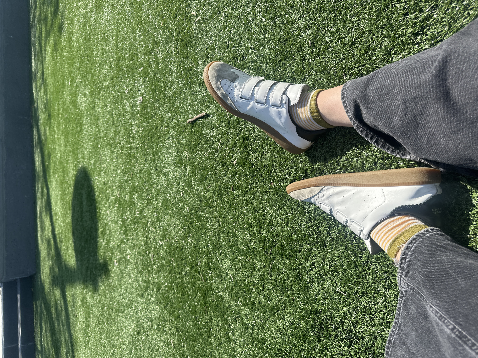 The image shows a person's legs and feet resting on a green, grassy surface. The person is wearing dark gray jeans and a pair of white sneakers with brown soles and velcro straps, along with striped socks. The grass appears to be well-maintained, possibly artificial turf. There is a shadow cast onto the grass, suggesting a sunny day. A small stick is visible near the shoe. The environment appears to be a park or recreational area, but the specific location within New York City cannot be determined from the image.