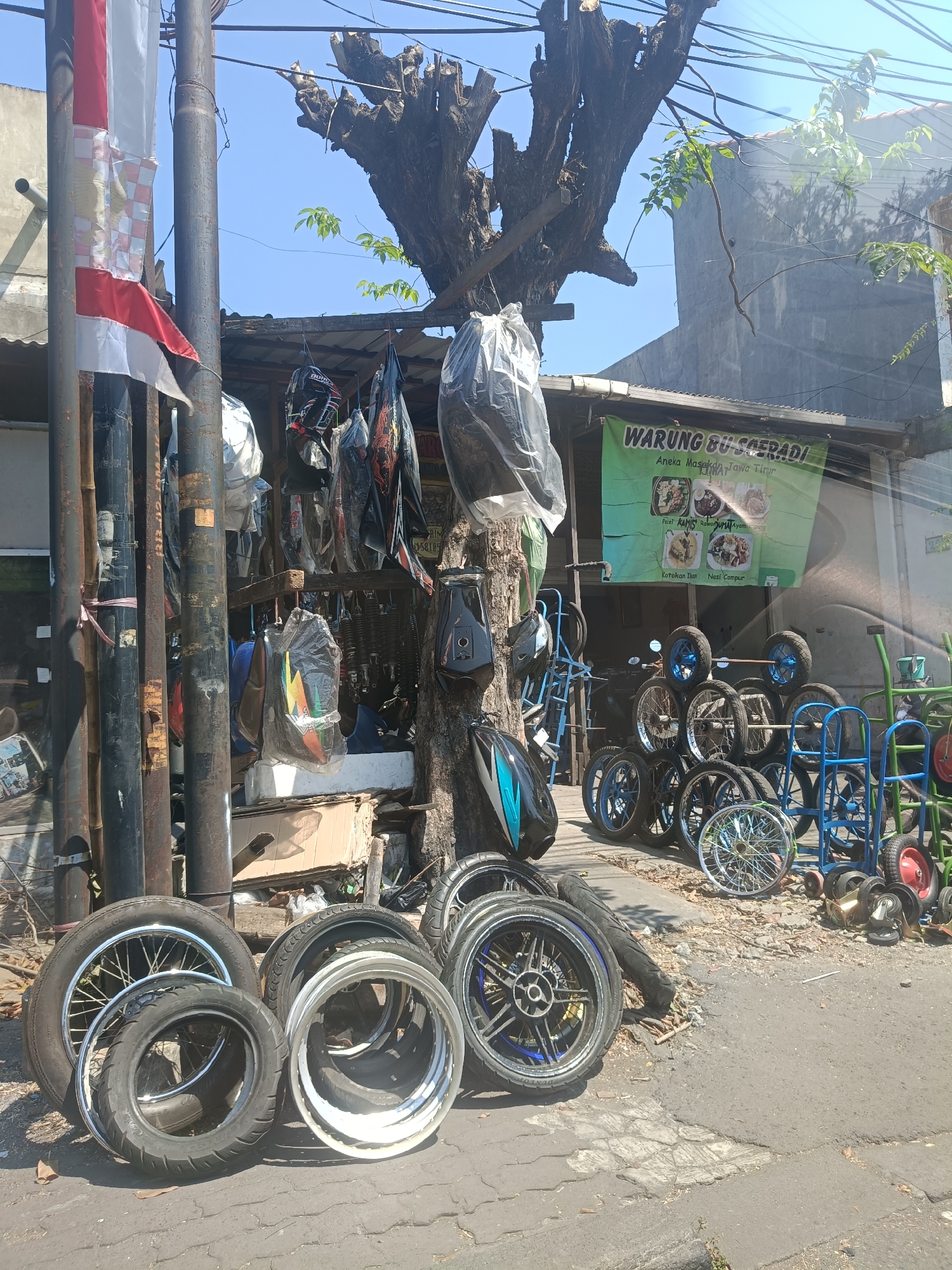 The image depicts a street-side stall in Surabaya, Indonesia, likely a motorcycle parts shop, situated beneath a large, dead tree. The stall displays a variety of items, including motorcycle wheels, tires, helmets, and other accessories. A banner for "WARUNG BU SOERADI" is visible, advertising local food specialties. Several metal poles, some bearing what appear to be national flags, are present, suggesting a public area. The overall scene is one of commerce and daily life in an urban Indonesian setting, with a focus on motorcycle-related goods.