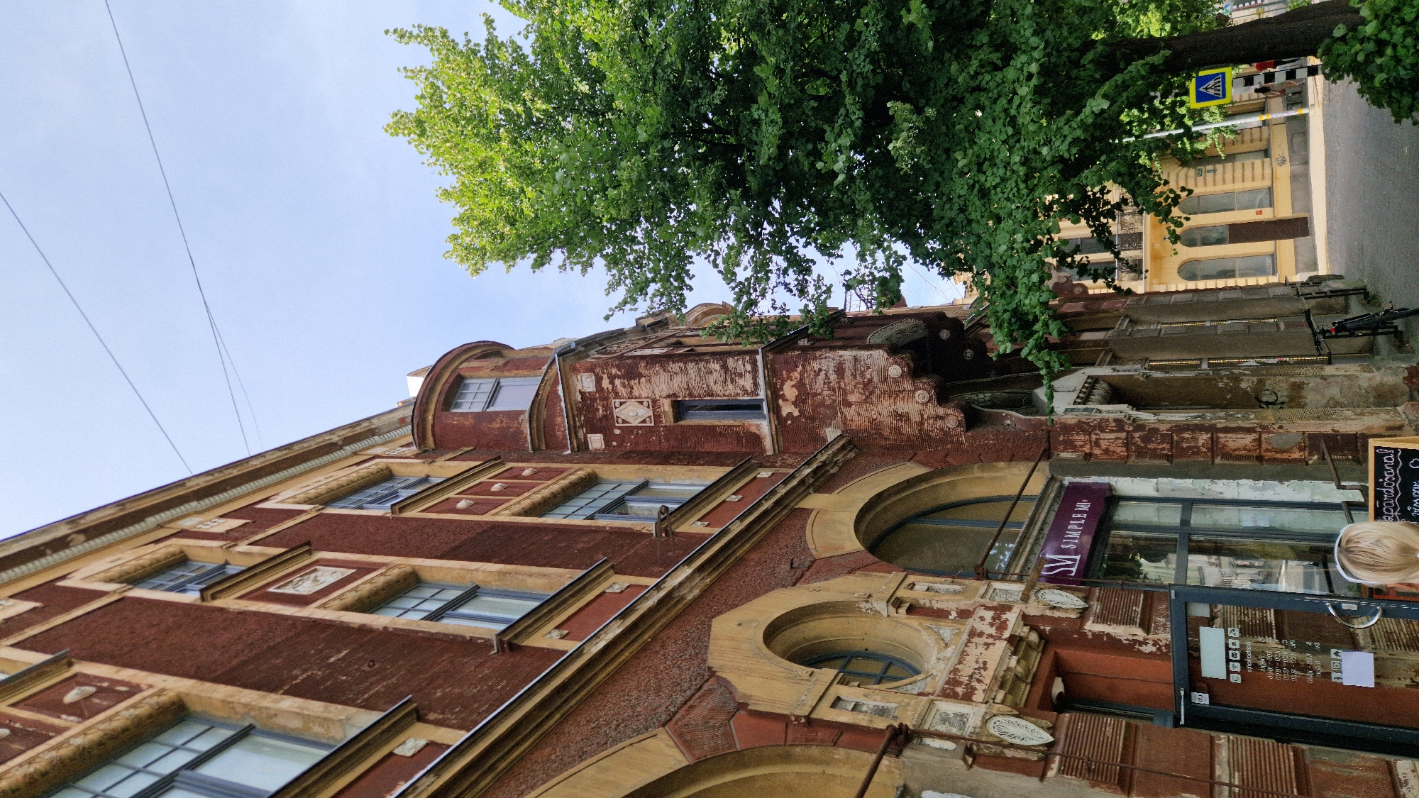 The image captures a street scene in Liepaja, Latvia, focusing on the facade of a multi-story building with distinctive red brickwork and yellow decorative accents. The building exhibits signs of age and weathering, with peeling paint and exposed plaster in some areas. Architectural features include arched windows on the lower levels and a rounded turret-like section on the upper right. To the right of the building, a large, leafy green tree dominates the frame, its branches reaching upwards. A sidewalk runs along the front of the building, with a bicycle parked near the entrance. A street sign with a pedestrian crossing symbol is visible further down the sidewalk, indicating a crosswalk. In the foreground, a person with blonde hair is partially visible, facing away from the camera. The sky above is clear and blue.