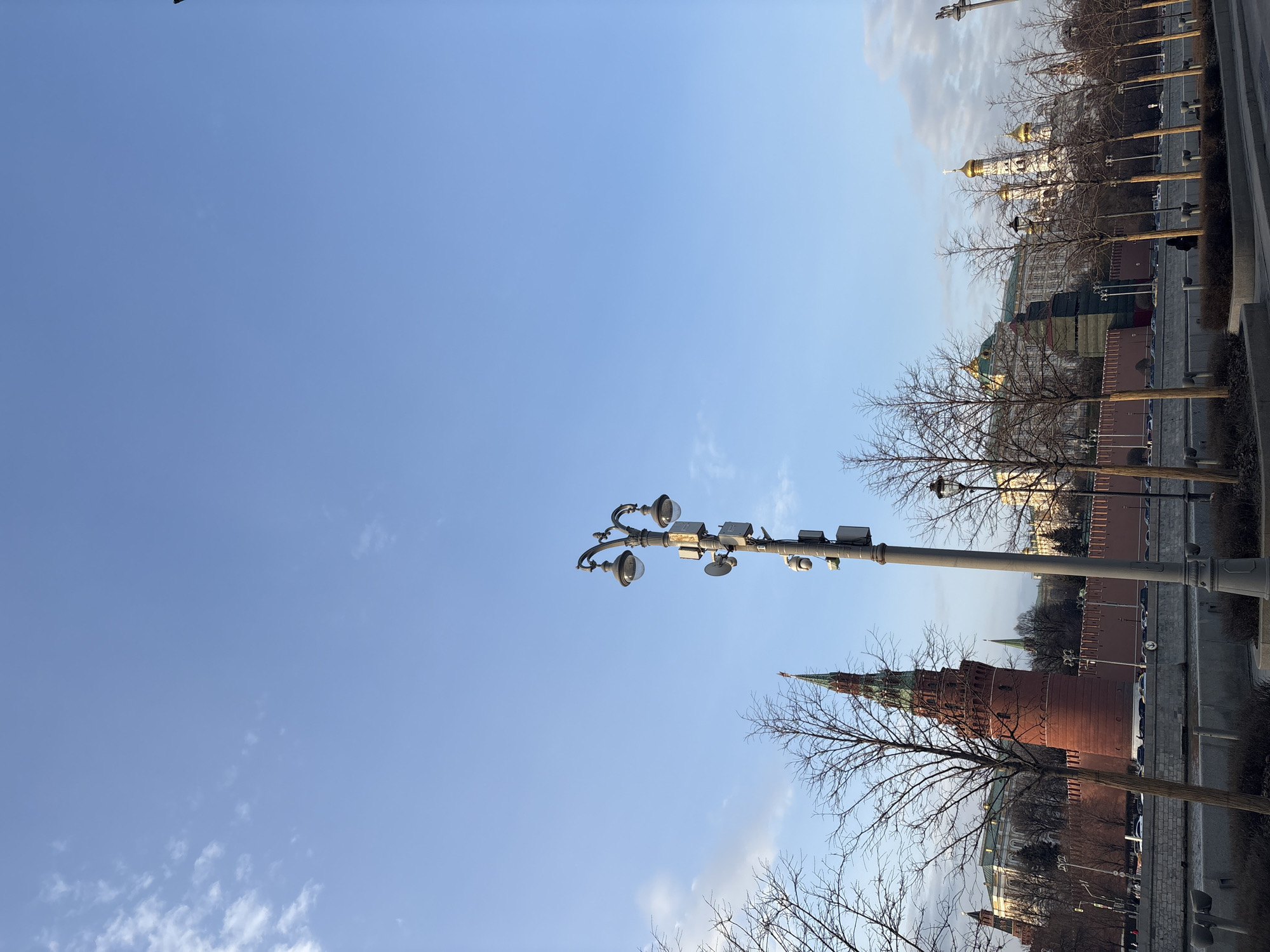 The image captures a view of the Moscow Kremlin in Russia. A lamppost with multiple lights and attached electronic devices stands in the foreground against a clear blue sky. The brick walls and towers of the Kremlin are visible, along with golden domes of cathedrals within the complex. Bare trees line the street beside the Kremlin wall. The scene suggests it is daytime, likely late autumn or early spring, given the leafless trees.