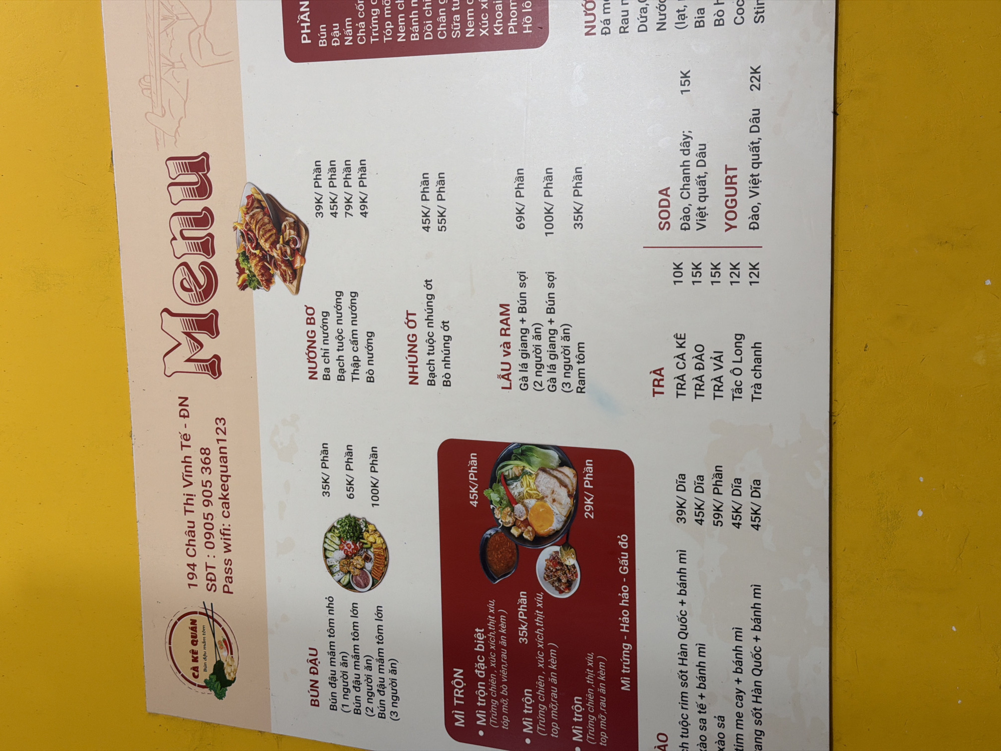 The image is a menu from a Vietnamese restaurant called "CÀ KẾ QUÁN" (Bún đậu mắm tôm), located at 194 Châu Thị Vĩnh Tế - ĐN in the city of Đà Nẵng, Vietnam. The menu is printed on a laminated surface, featuring a variety of Vietnamese dishes, including bún đậu (fermented shrimp paste noodles), grilled items (nướng bơ), dishes with nhúng ớt (dishes with chili), hot pot and ram (lẩu và ram), and mì trộn (mixed noodles). The menu also lists various drinks such as trà (tea), soda, and yogurt. Prices are listed in Vietnamese currency (VND). The restaurant's phone number and wifi password are also visible on the menu. The image is cropped and shows only a portion of the menu.