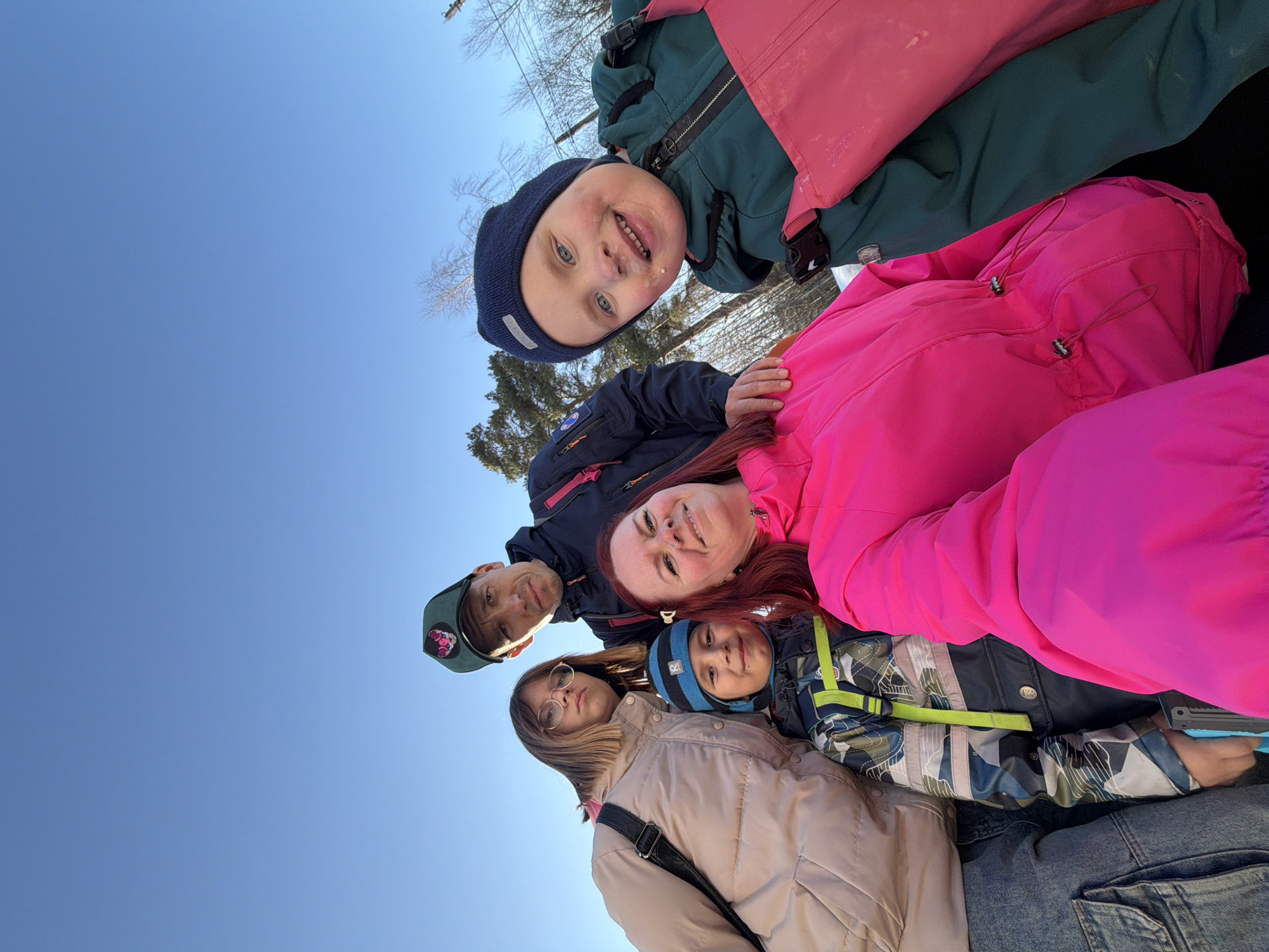A group of six people are standing close together, looking up at the camera, which is positioned at a low angle against a clear blue sky. The group includes two younger children wearing winter hats and brightly colored jackets (one in pink and one in green), two older children in winter clothing, and two adults. They are outdoors, and some bare tree branches are visible in the upper background. The precise location in the city of Chaikovsky, Russia, cannot be determined from the image.