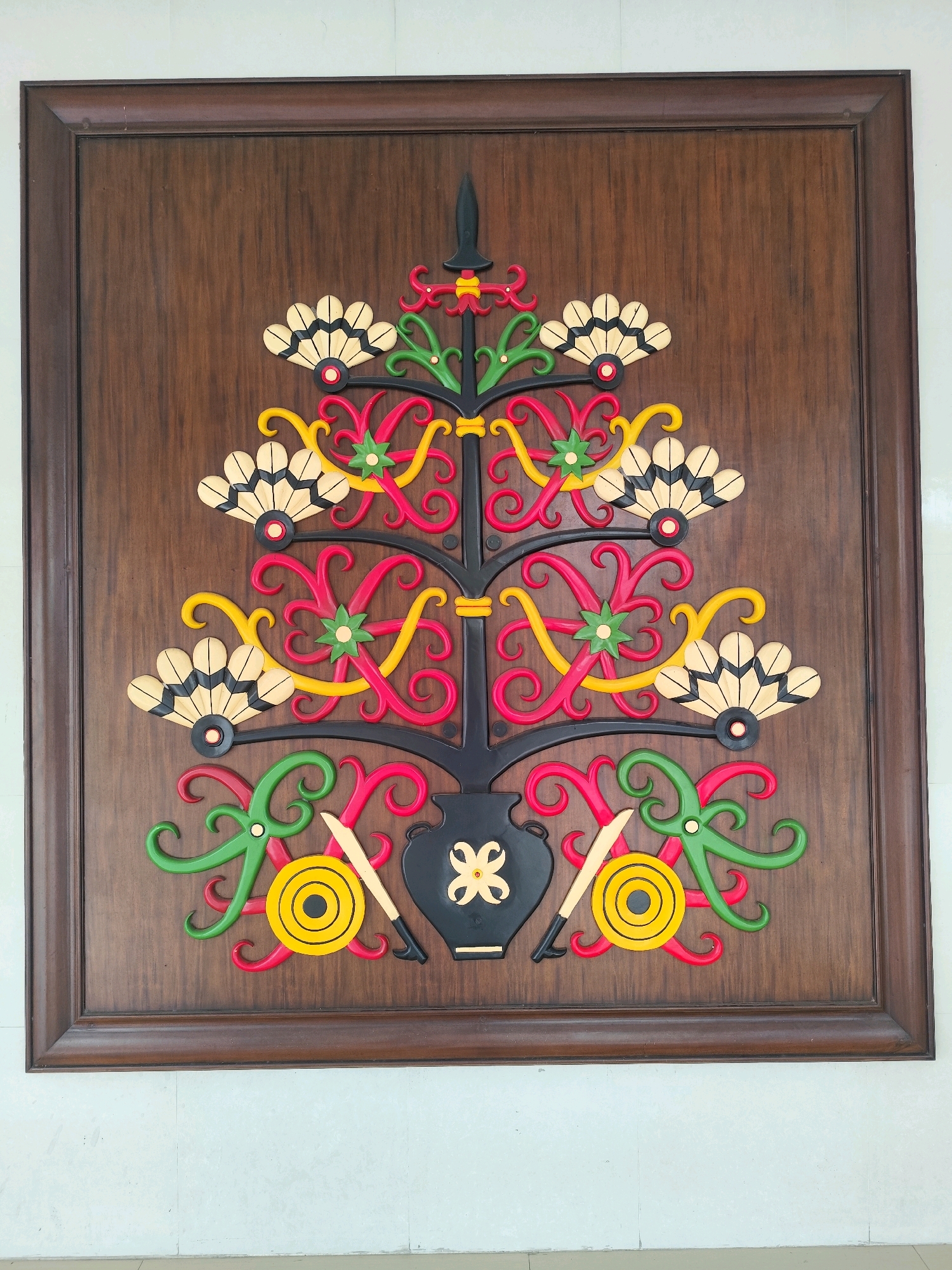 The image shows a carved wooden panel featuring a colorful, abstract tree-like motif. The panel is framed by a dark brown wooden border and is mounted on a wooden surface. The carving is intricate, with swirling shapes in red, yellow, and green, and fan-shaped elements in beige and black. At the base of the motif is a black vase-like object with a yellow flower design. Two circular yellow objects with black centers and two light-colored swords with black handles are also depicted at the bottom. The overall style suggests a cultural or traditional art form. This artwork is located at the Betang Bawi, a traditional Dayak house in Palangka Raya, Indonesia, which serves as a cultural center and museum.