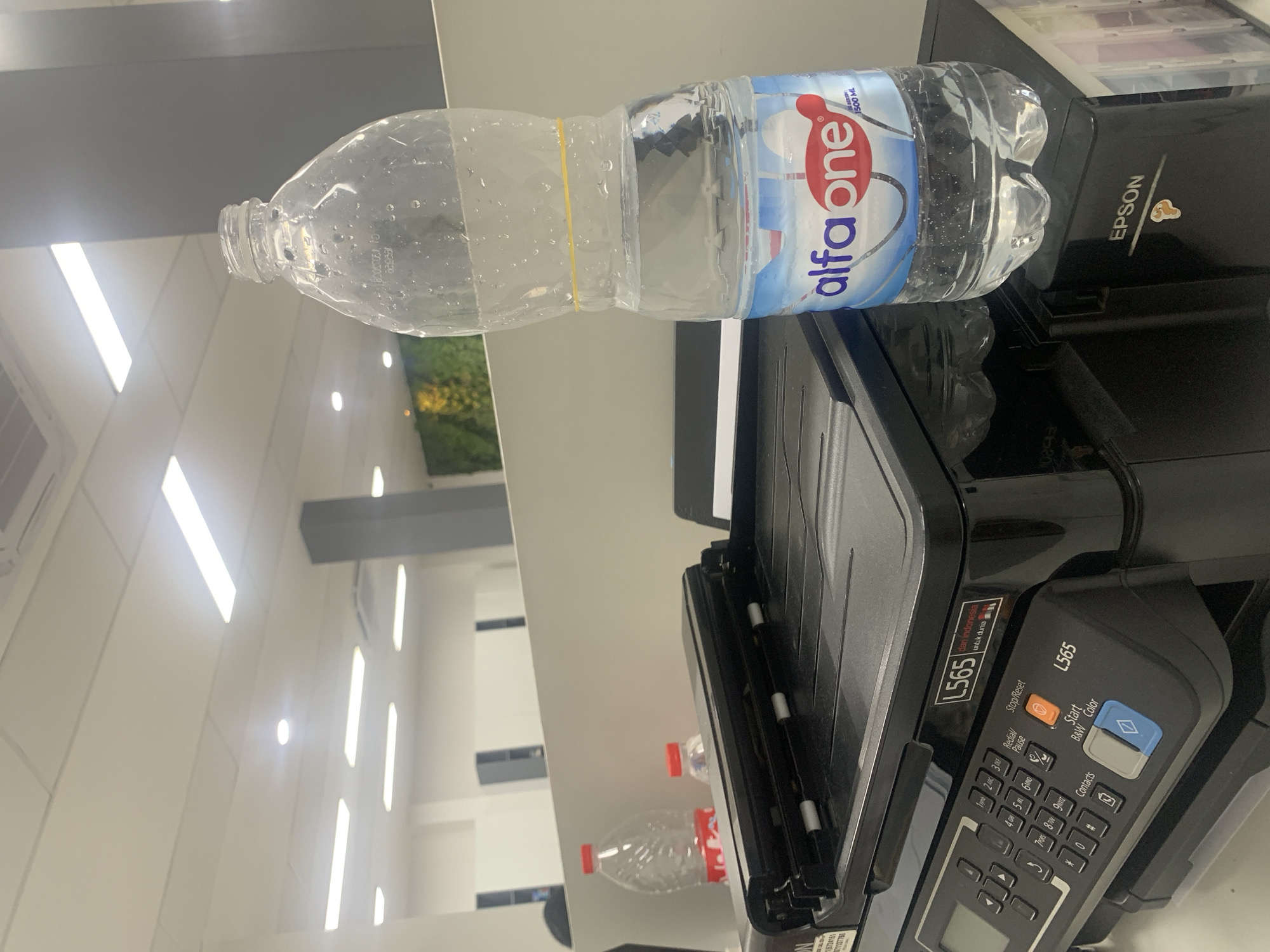 In an office environment in Palembang, Indonesia, a transparent plastic water bottle from "alfa one" with a capacity of 1500 ML is placed on top of a black Epson L565 printer. The bottle contains water and has a yellow rubber band around its neck. The printer is positioned on a white desk, and the background reveals a modern office setting with ceiling lights and a blurred partition wall. There is another red-capped water bottle partially visible to the left of the main subject. The scene is brightly lit, suggesting an indoor commercial or administrative space.