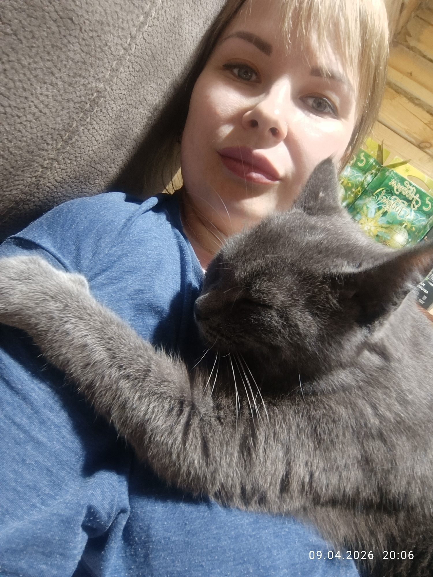 A woman with light blonde hair is holding a gray cat with its eyes closed. The cat is nestled on the woman's blue shirt and has its paw around her shoulder. The background includes a gray couch and a green box with text in Russian. The image includes a timestamp indicating it was taken on April 9, 2026, at 20:06. The location is presumed to be in Chaikovsky, Russia.