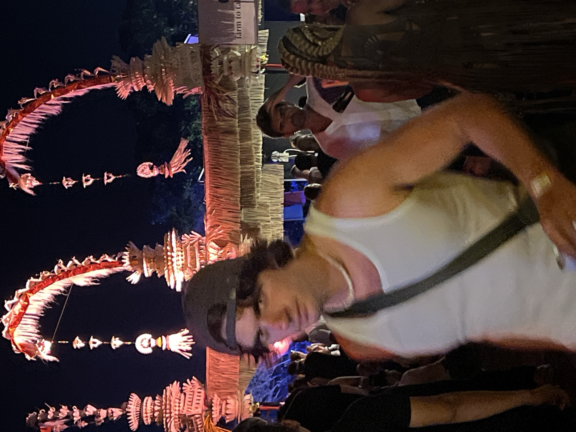 The original image depicts a nighttime scene at what appears to be a lively outdoor festival or cultural event. The perspective is tilted, with subjects and structures oriented vertically on the left and right, suggesting the camera was held at an angle.
In the immediate foreground, a person is visible, slightly blurred by motion. They are wearing a white tank top and a dark baseball cap, looking towards the viewer. A dark strap, possibly from a bag, crosses their chest. Their arm is partially visible on the right.
Behind this person, another individual wearing a white top is smiling, their face clearly visible. They have distinct braided hair or a textured head covering. Further into the background, numerous indistinct figures suggest a significant crowd gathered for the event.
The setting is characterized by elaborate, illuminated decorations. On the left, two large, arching structures, resembling skeletal dragons or ornate garlands, curve upwards. These are made from natural materials, possibly straw or wood, and glow with a warm, reddish-orange light. Below these arches, smaller, intricate decorative elements hang suspended. To the right, a tall, vertical pillar made of woven material, possibly bamboo or straw, stands prominently. Attached to this pillar is a light-colored sign fragment that reads "farm to ca," likely part of "farm to table."
The dim overall lighting indicates it is nighttime, with artificial lights highlighting the decorative elements and subjects. The scene conveys a festive and engaging atmosphere.