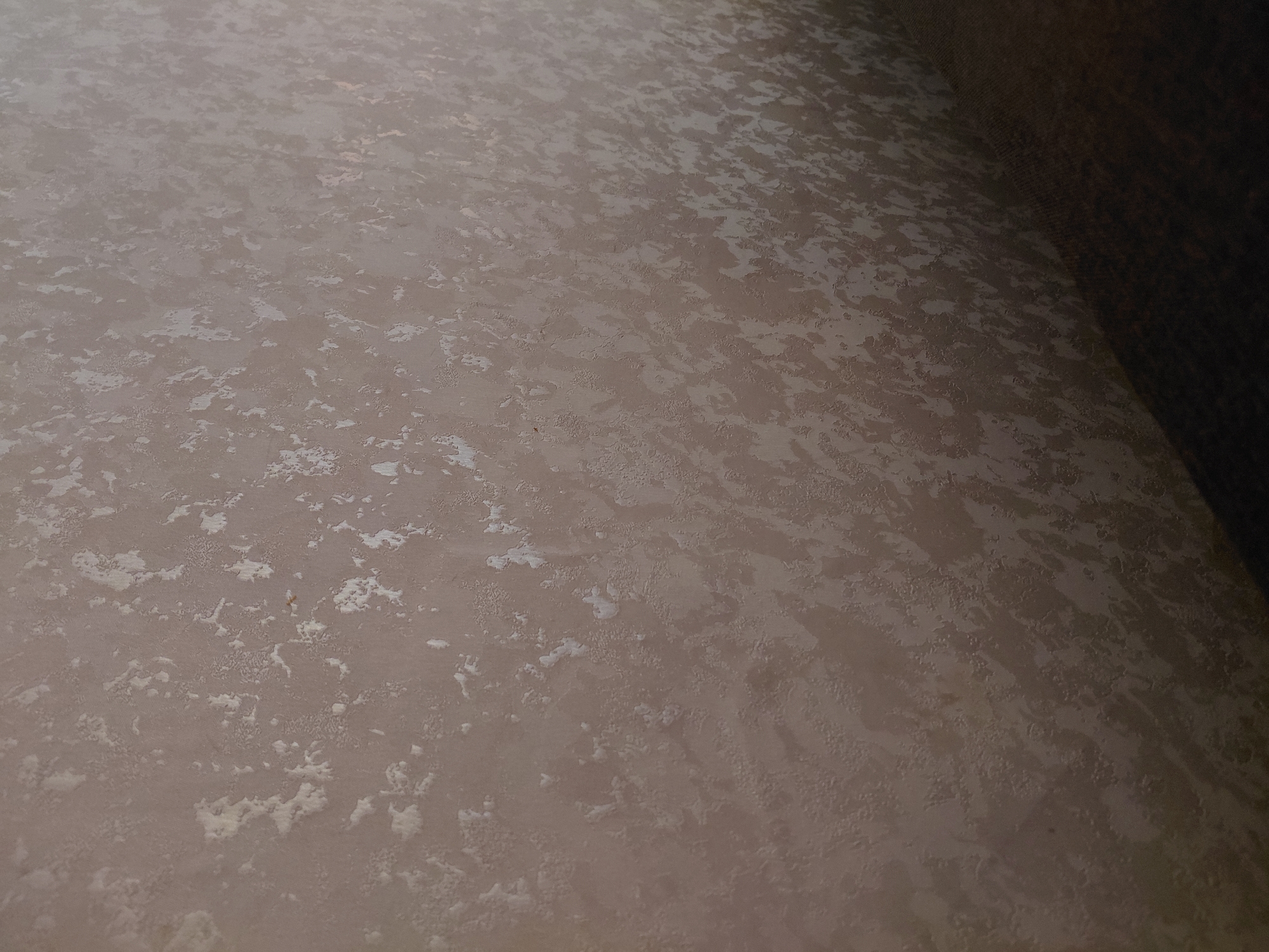 The image shows a close-up, textured surface, possibly a wall or fabric, with a mottled pattern of light and dark areas. The texture appears to be raised in some places, creating a slightly irregular, almost stucco-like appearance. The dominant color is a neutral beige or taupe, with lighter, almost white patches scattered across the surface. The lighting casts subtle shadows, highlighting the dimensionality of the texture. The lower right portion of the image shows a darker, undefined area, possibly furniture or a shadow. There are no discernible objects, people, or actions taking place. The location in Malinovka, Russia, is not visually represented in the image.