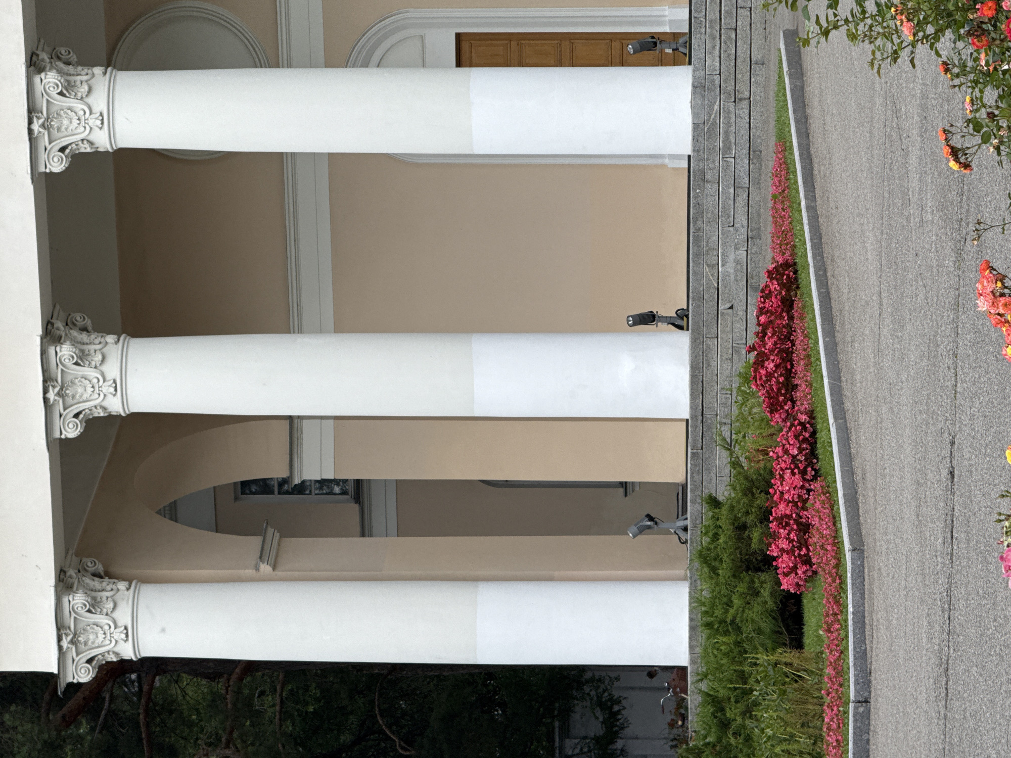 The image shows the portico of a building, characterized by three large white columns with ornate capitals. The building's facade is painted a pale peach color, with arched openings and a wooden door visible. In front of the building, a tiered stone staircase leads up to the entrance. A bed of vibrant pink flowers borders the staircase and the paved walkway. Two small, dark spotlights with stands are positioned on the stone steps. To the left of the building, a dark green evergreen bush is visible, and further left, a glimpse of a bicycle and possibly a person can be seen against a dark background of trees. The foreground consists of a gray asphalt road, with a section of blooming roses in the lower right corner. The scene depicts the exterior of a building, likely a public or institutional one, in a well-maintained garden setting. The style of the architecture suggests it could be a historical or neoclassical structure. The lighting indicates it is daytime.