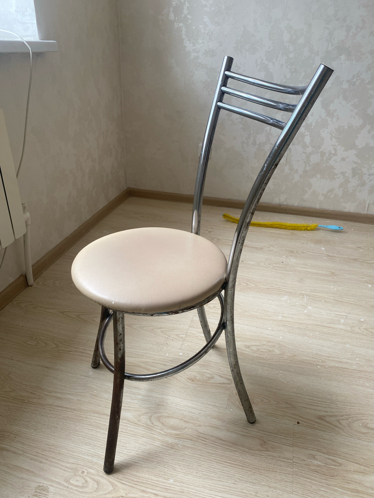 A metal stool with a light-colored, cushioned seat stands on a light-colored wooden floor. The stool has a curved metal backrest with three horizontal bars. The metal frame appears aged and somewhat rusted in places. To the right of the stool, on the floor, lies a yellow duster with a blue handle. The background consists of a textured, light-colored wall. To the left, part of a white radiator or baseboard is visible near a window with light coming through. The scene appears to be indoors, possibly in a room in Oyskhara, Russia, given the context. There are no people or animals present. The lighting is natural and appears to be coming from the window.