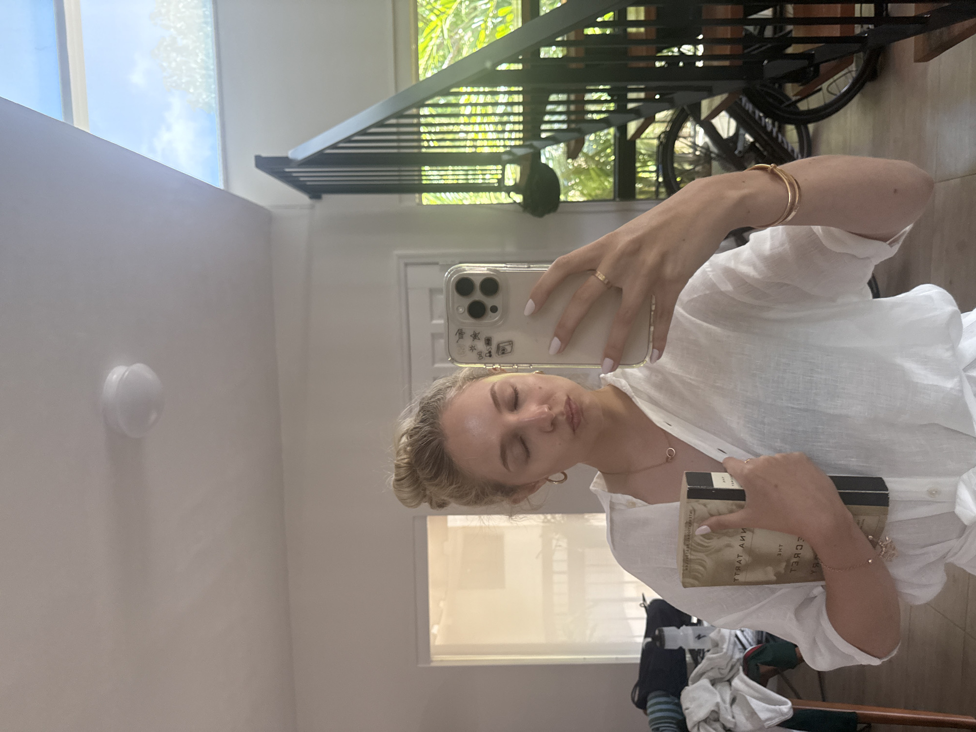 A woman with her hair in a bun is taking a selfie in a mirror. She is wearing a white linen shirt and holding a book titled "The Secret" by Rhonda Byrne. She has on jewelry including a necklace, earrings, and bracelets. In the background, there is a staircase and a skylight with a view of trees and the sky. The scene appears to be indoors, likely in a residential setting. There is no indication of the city of Miami, United States of America.