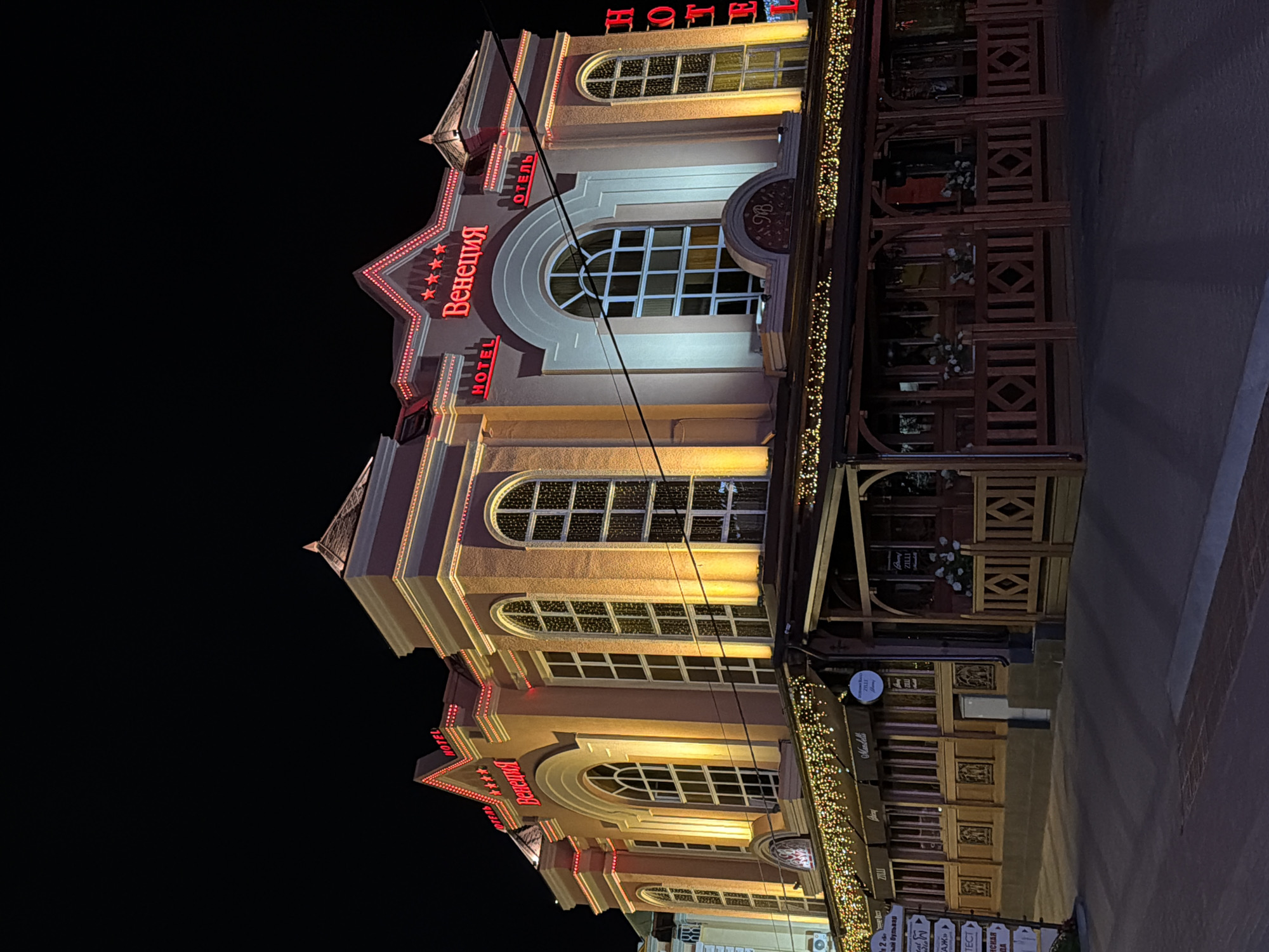 The image shows a brightly lit hotel named "Венеция" (Venetsiya), meaning Venice, at night in Kislovodsk, Russia. The hotel features ornate architecture with a stucco facade, arched windows, and decorative red neon lights spelling out "HOTEL" and the hotel's name. String lights also adorn the exterior, illuminating the building and the outdoor seating area of a restaurant or cafe below. The scene is composed of the hotel's facade, its illuminated signage, and the entrance to a commercial establishment on the ground floor. The surrounding environment is dark, indicating it is nighttime, with hints of a street or sidewalk in the foreground. No people are visible in the image.