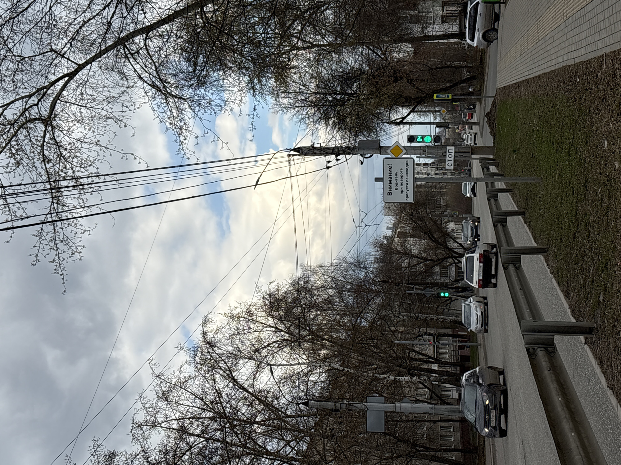 The image depicts an overcast daytime street scene in what appears to be late autumn or early spring, given the bare branches of the deciduous trees lining the road. The perspective is from the roadside, looking down a multi-lane road with a concrete barrier or guardrail separating it from a grassy strip.
Several cars are visible on the road, including a dark-colored sedan in the foreground, a silver car, and a white car further down the road, and another white car on the right side of the road, parked or driving on a side street or entrance. Traffic lights are visible, with the green signal illuminated, suggesting vehicles may be waiting at an intersection or moving through one.
Utility poles with numerous overhead wires crisscross the sky, adding to the urban landscape. A pedestrian sidewalk runs parallel to the road on the right.
Notable details include a traffic light pole with several signs. A prominent white rectangular sign displays Cyrillic text: "Внимание! Водитель, при повороте пропусти пешеходов", which translates to "Attention! Driver, when turning, yield to pedestrians." Below this, a small "СТОП" (STOP) sign is visible. Above the yield sign, a yellow diamond sign, typically indicating the main road, is present. The leafless trees and the general light suggest a cool, possibly early spring or late autumn day in Izhevsk, Russia. No people are explicitly visible in the frame. The overall mood is functional and everyday urban.