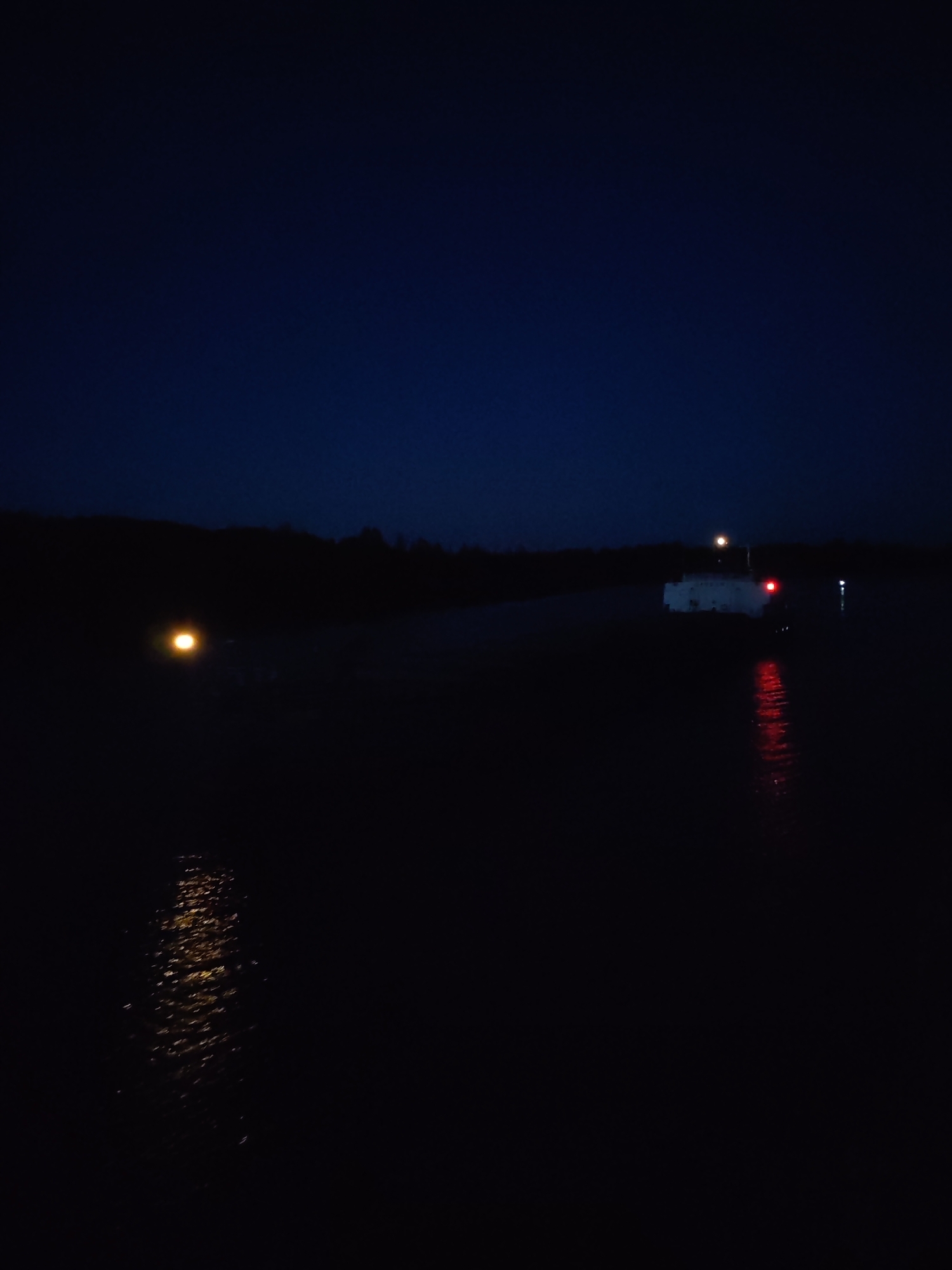 At night in Badozhskoi Pogost, Russia, a body of water reflects light from visible structures and points of light. In the middle ground, a white structure with a bright light on top and a red light on the side stands on what appears to be land. To the left, a single orange light is reflected in the water. Trees are visible on the horizon.