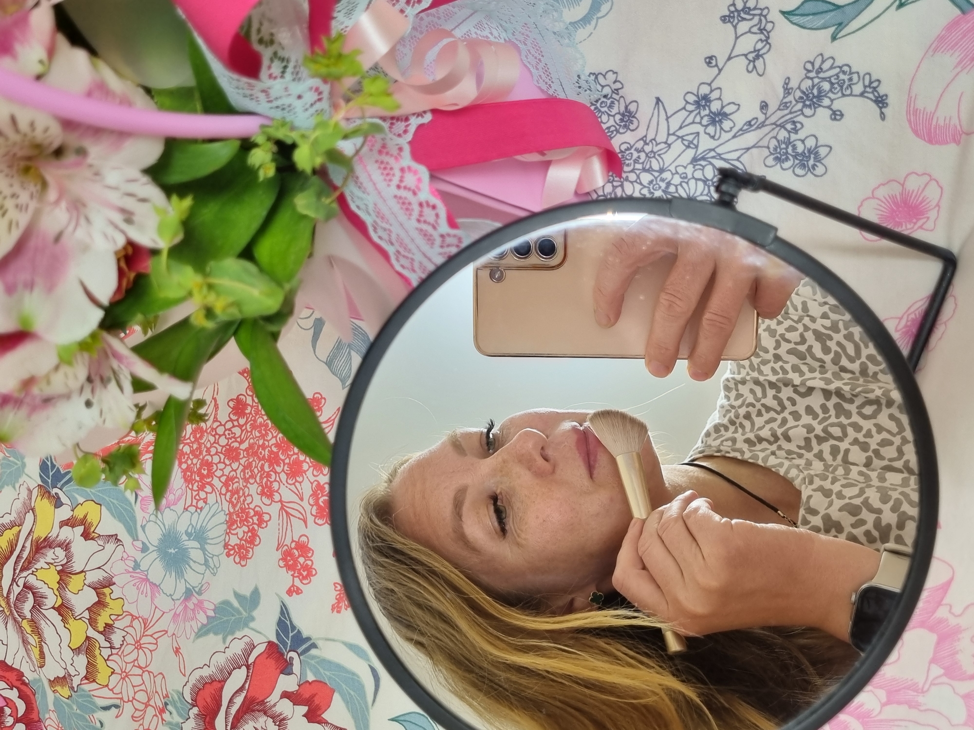 A round mirror with a black frame is centrally positioned, reflecting a woman engaged in applying makeup. The woman, with light brown hair and visible freckles, holds a light gold-colored smartphone in her left hand, positioned as if taking a selfie. In her right hand, she holds a makeup brush with a golden handle, touching it to her face near her upper lip. She wears a light-colored top with a subtle leopard print and a smartwatch on her left wrist.
Outside the mirror's reflection, the background is dominated by a vibrantly patterned fabric, likely a bedsheet or tablecloth, adorned with large floral designs in shades of red, yellow, blue, and pink against a white base. To the upper left, a bouquet of pink and white alstroemeria flowers with green foliage is partially visible, embellished with bright pink and light blue lace ribbons.
The scene is brightly lit, suggesting natural daylight in an indoor setting, possibly a bedroom or dressing area, given the personal grooming activity and domestic textiles. The woman's expression appears focused and neutral. There is no discernible text visible in the image.