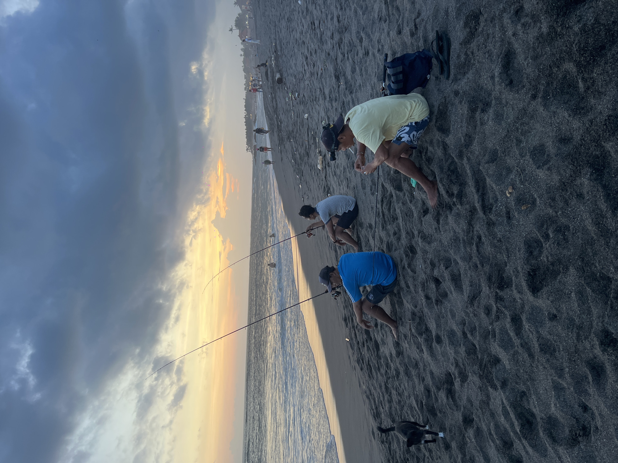 Three men are fishing on a black sand beach in Mengwi, Indonesia. They are crouched near the shoreline, each holding a fishing rod. A dark-colored dog is standing near one of the men. There are people walking along the beach further in the background. The sky is overcast with a sunset glow on the horizon. A backpack and a pair of sandals are on the sand. The beach has some trash.