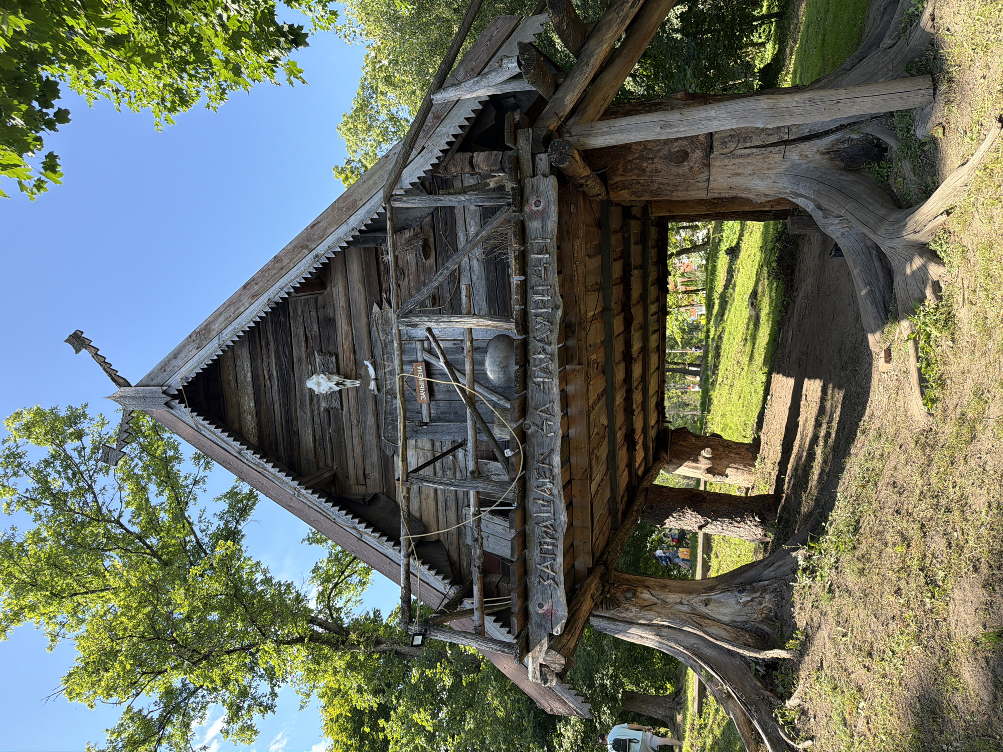 The image depicts a rustic, wooden structure resembling a hut or a small house, elevated on thick, gnarled tree stumps. The structure has a steep, gabled roof with decorative wooden trim along the eaves. A balcony or porch area wraps around the front of the structure, supported by additional wooden beams. The facade of the hut is adorned with a skull, possibly of an animal, and various wooden carvings and inscriptions. A large, dark, round object, possibly a pot or a cauldron, hangs from the balcony. The hut is situated in a grassy, park-like environment with several large, mature trees providing shade. Sunlight dapples the ground, creating shadows beneath the structure. In the background, there are glimpses of other people walking and trees. The inscription on the front of the structure, visible from the ground, appears to be in Cyrillic script. This structure is identified as a place of interest in the Litvensky Selsoviet, Belarus, likely an attraction within a rural or historical park.