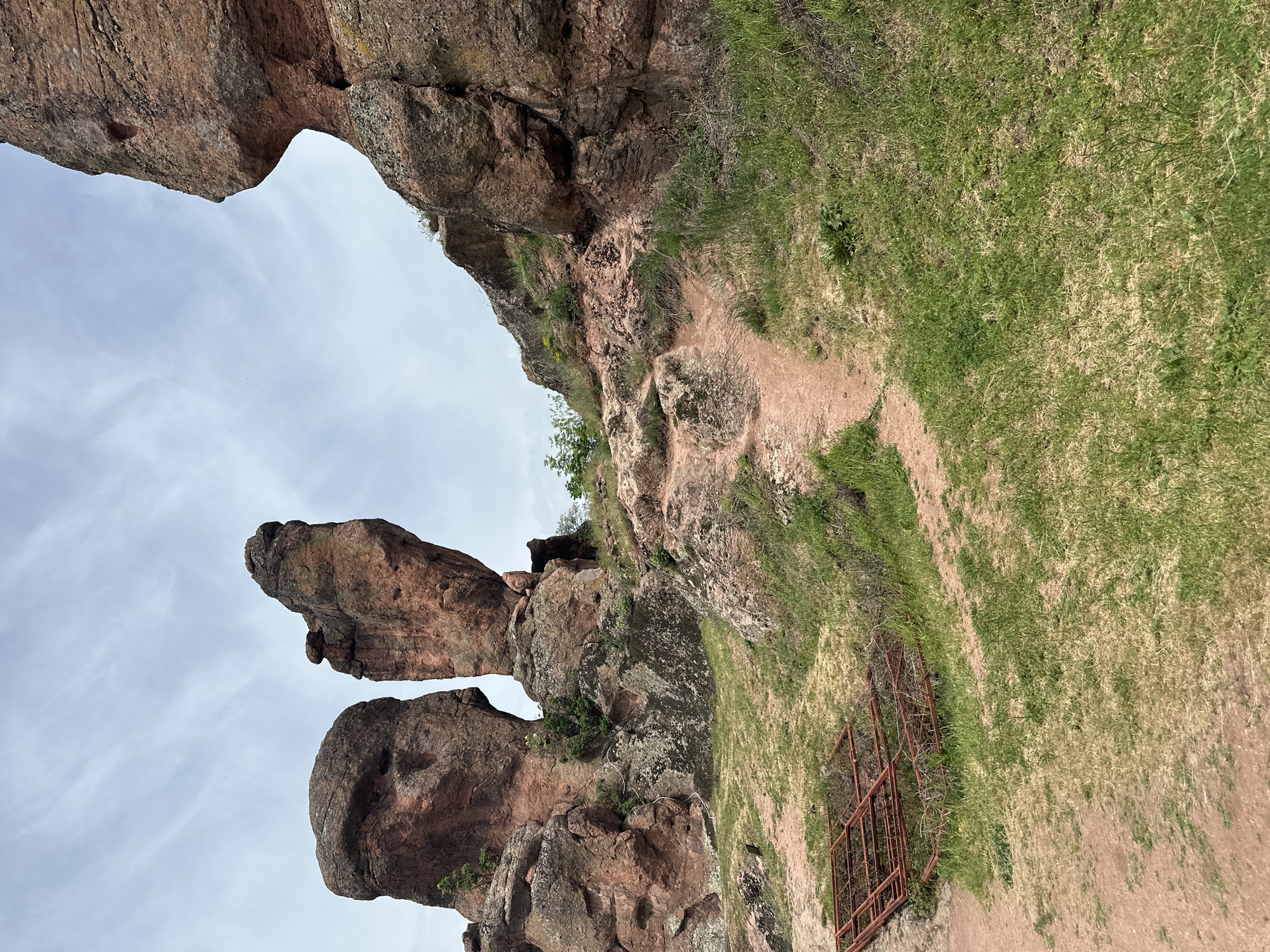 The image depicts a low-angle view of rock formations in Belogradchik, Bulgaria. The formations are primarily reddish-brown, weathered rock, and there is a small red metal fence near the ground. The sky is visible between the rock formations, with a partly cloudy sky. Patches of green grass are also visible near the rock formations.