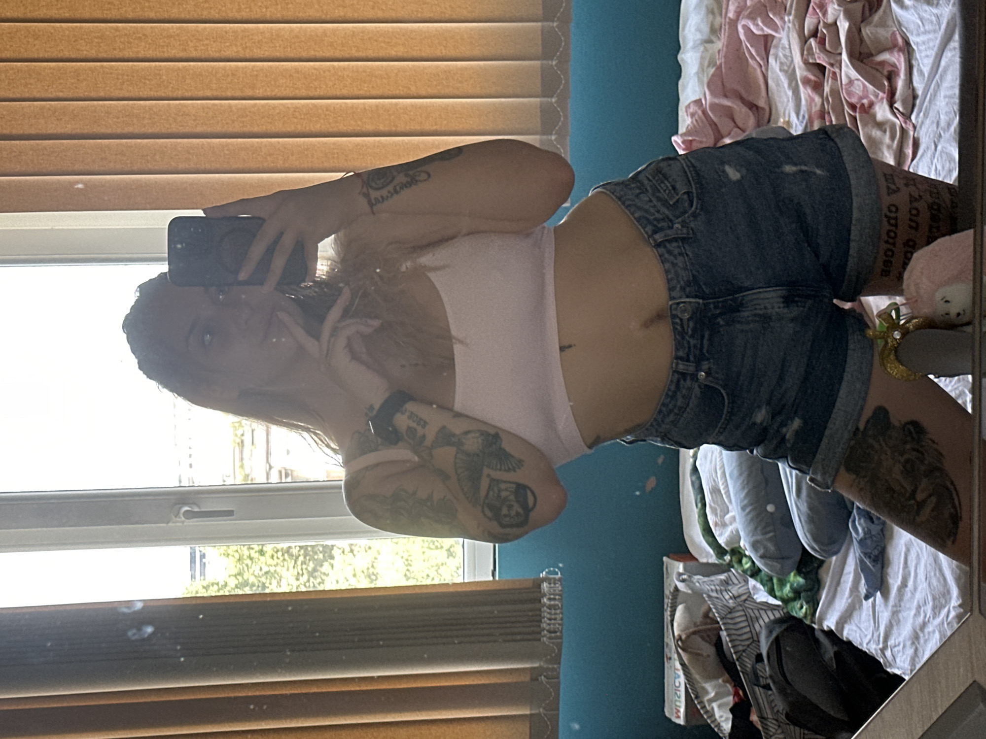 A woman with long blonde hair is taking a mirror selfie in a room. She is wearing a light pink crop top and denim shorts. She has multiple tattoos on her arms and leg. Her right hand is holding a black smartphone, and her left hand is near her face. The background shows a window with vertical blinds and a messy bed with various items, including pillows and clothing. The room appears to be a bedroom. The location is a bedroom in Plovdiv, Bulgaria.