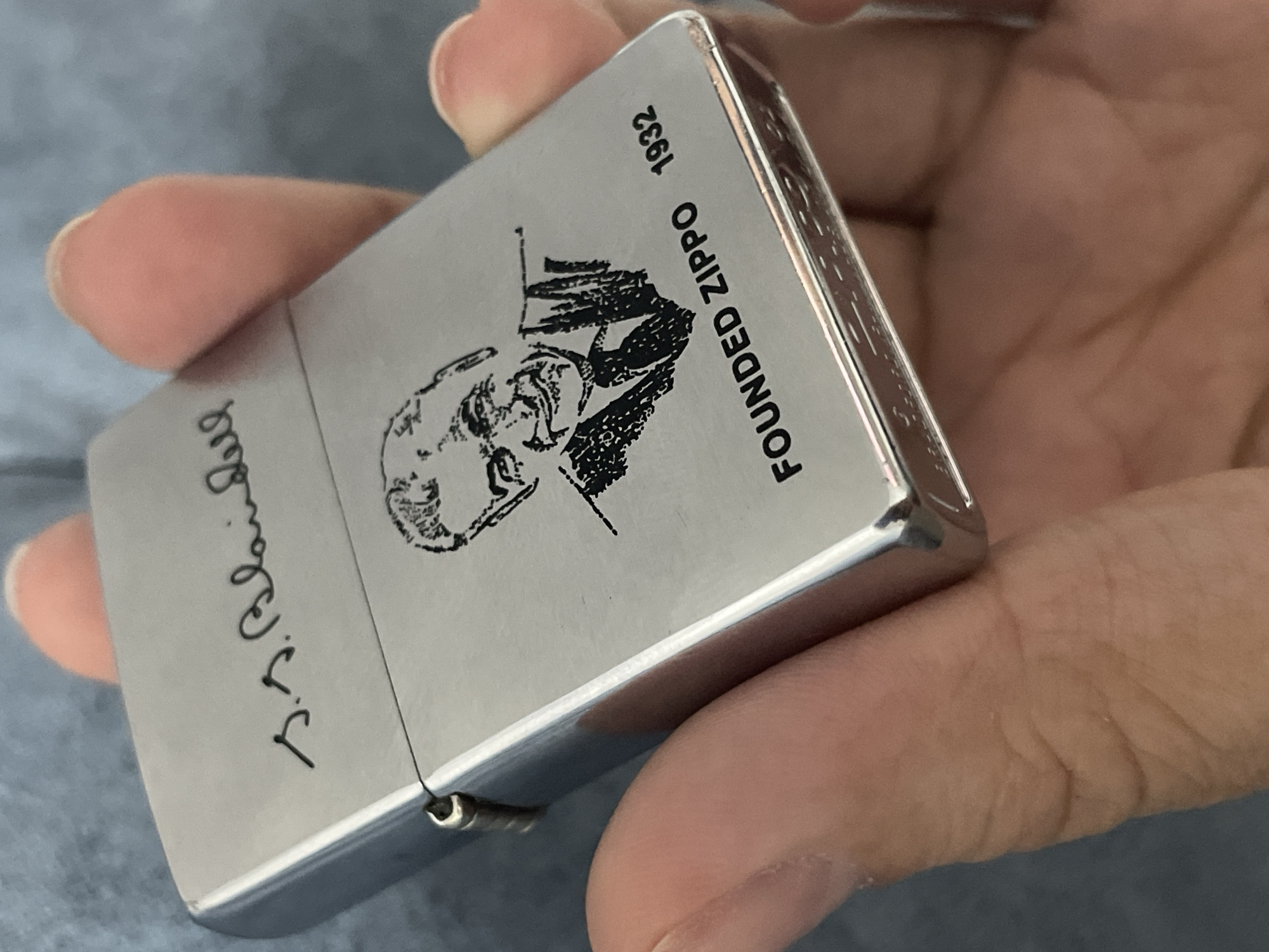 A person holds a silver Zippo lighter in their hand. The lighter has a black and white illustration of a man's face and the words "FOUNDED ZIPPO 1932" printed on one side. On the other side, there is a handwritten signature in black ink. The background is blurry and appears to be a neutral-colored fabric. There is no indication of the location being Izhevsk, Russia.