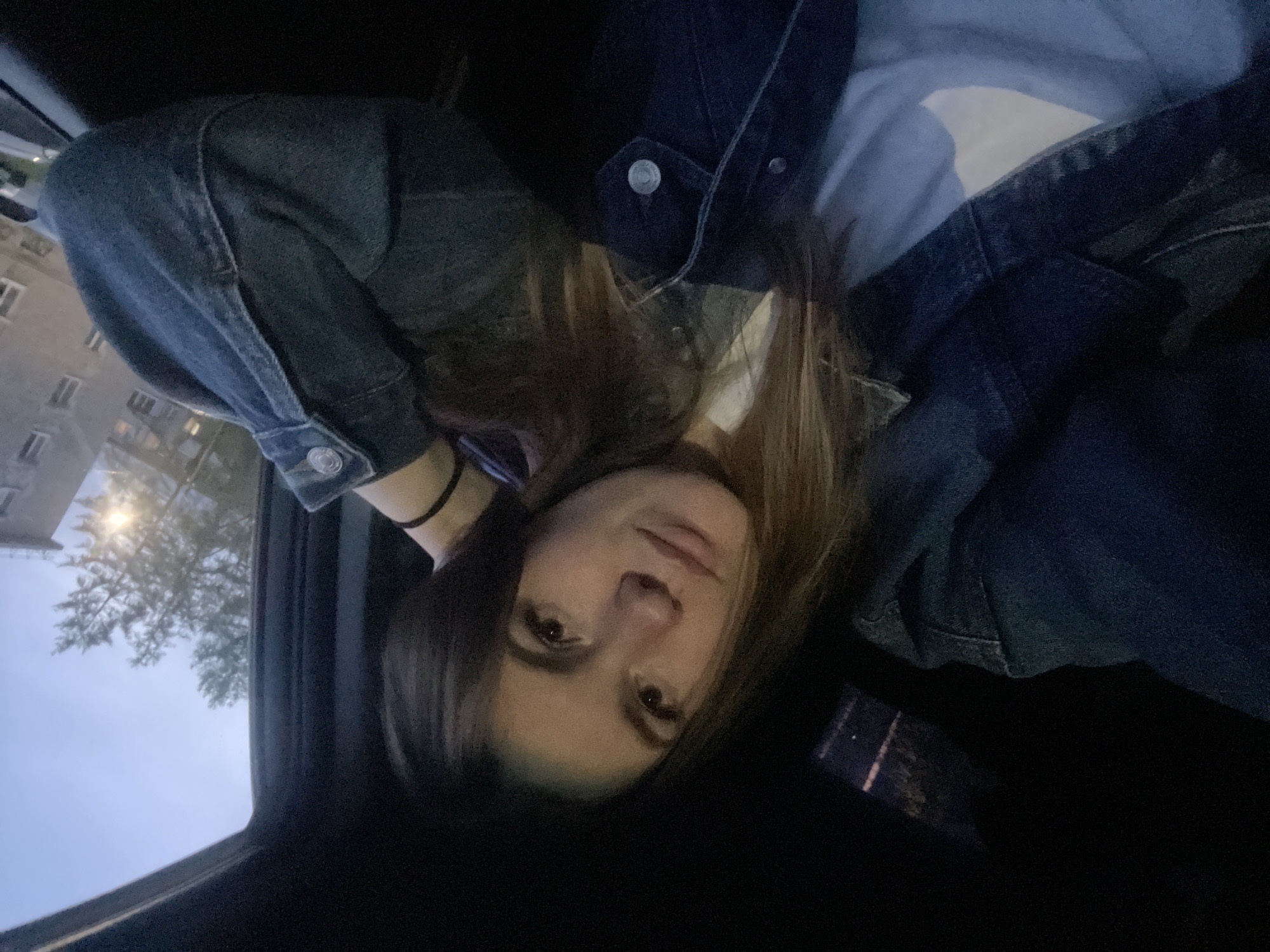A person is in what appears to be the front seat of a car. The person has long brown hair, brown eyes, and is wearing a blue denim jacket over a lighter-colored shirt. They have a black choker necklace. Outside the car window, there is a building and trees, suggesting an urban environment. The city is Chaikovsky, Russia.