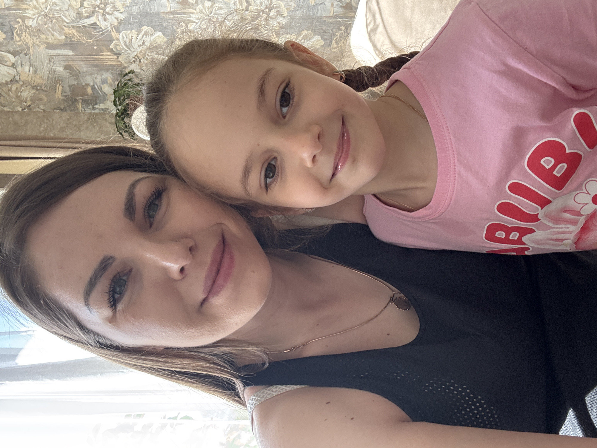 A woman and a young girl are posing for a selfie. The woman has light brown hair and is wearing a black tank top and a gold necklace. The girl has light brown hair in braids and is wearing a pink t-shirt with red and white graphics. They are both smiling at the camera. The background appears to be a room with patterned wallpaper and a sofa. The location is Раздолье, Russia.
